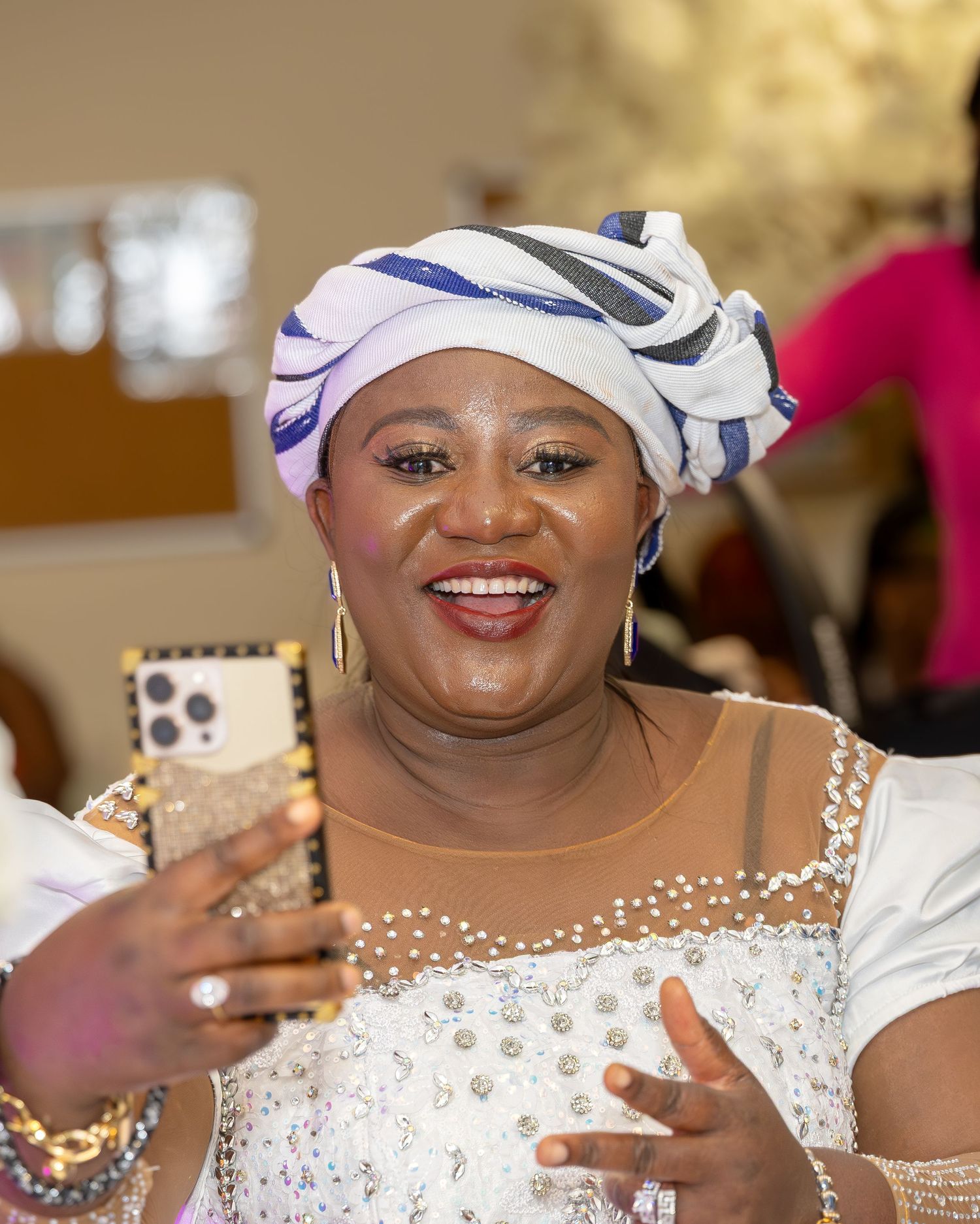 Joyful woman in white and blue gele headwrap taking selfie at Nigerian traditional wedding celebration.