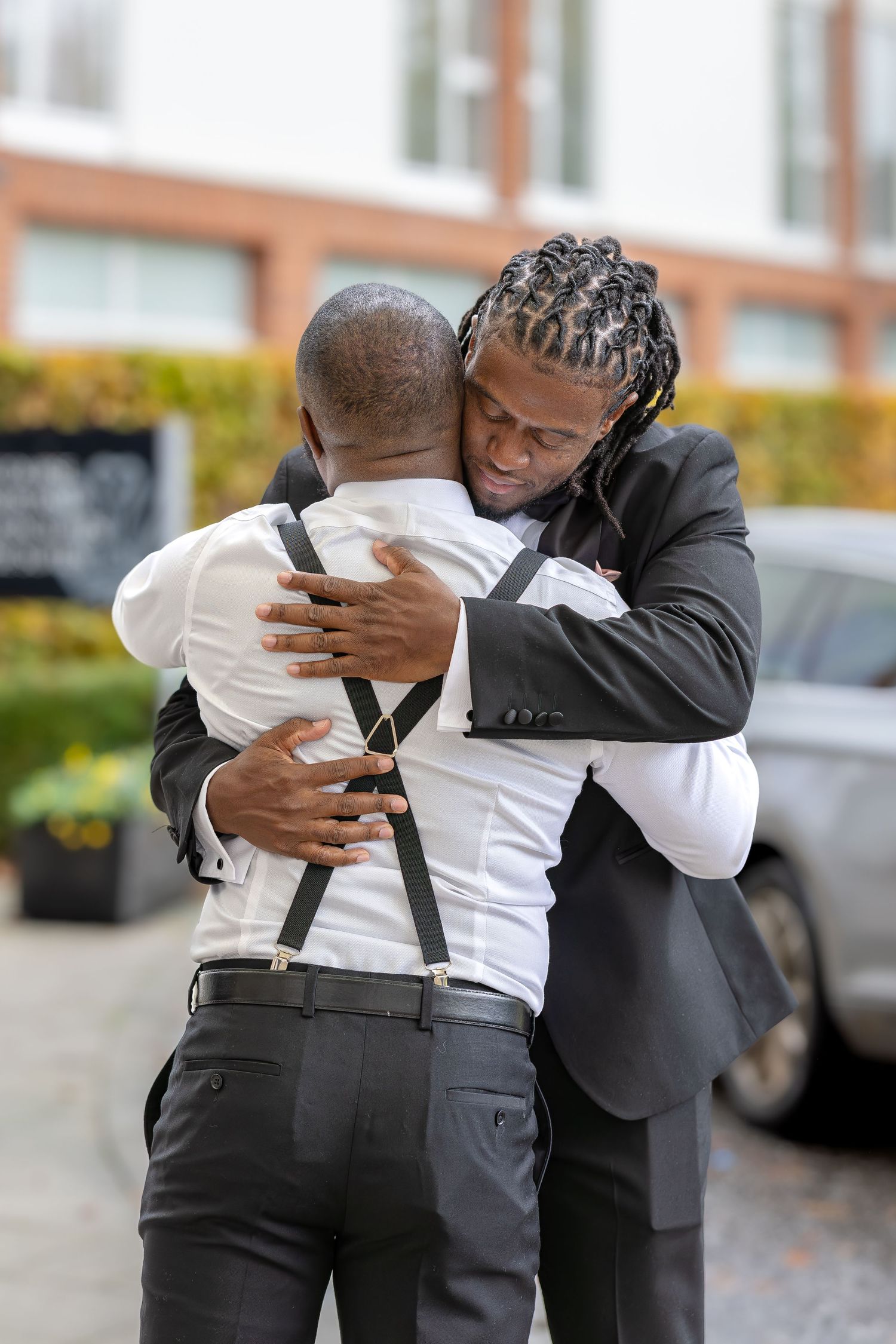 Two men in suits share an emotional hug outdoors at a wedding celebration.