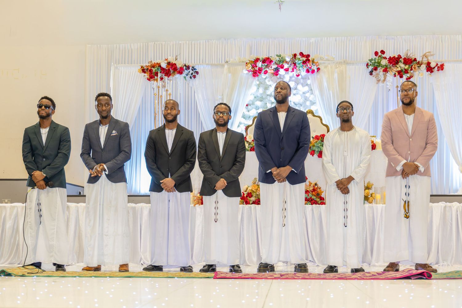 Seven men in traditional African attire stand in a row before a floral backdrop at a cultural ceremony.