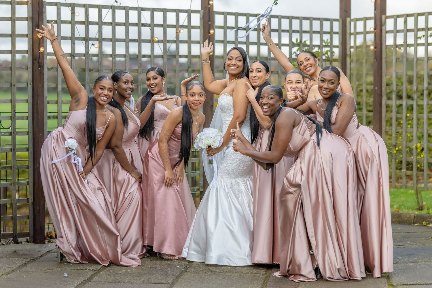Joyful bride and bridesmaids in pink satin gowns celebrate together with fun poses at an outdoor wedding venue.