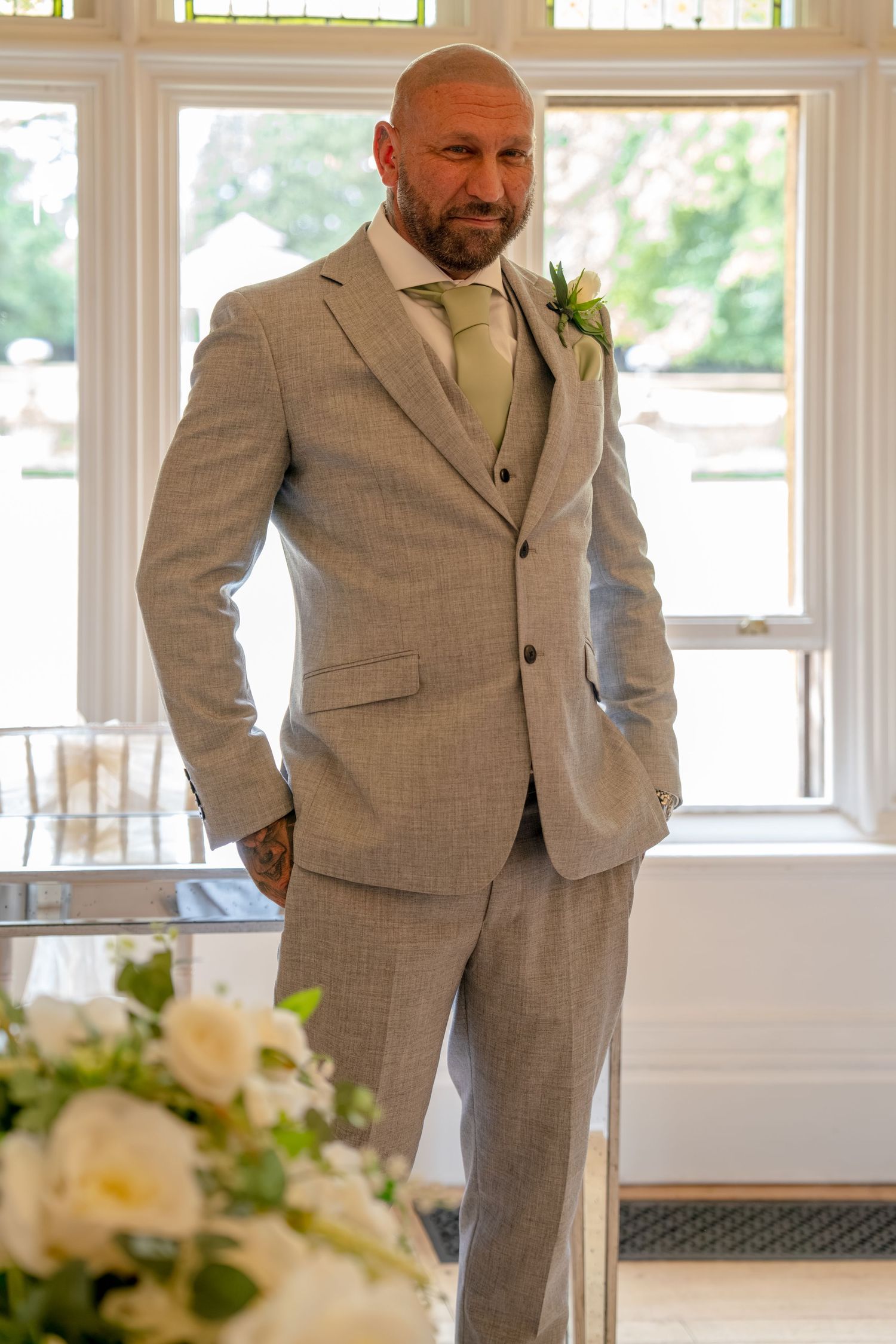 Bald groom in light gray three-piece suit with green tie and white boutonniere stands confidently at wedding venue.