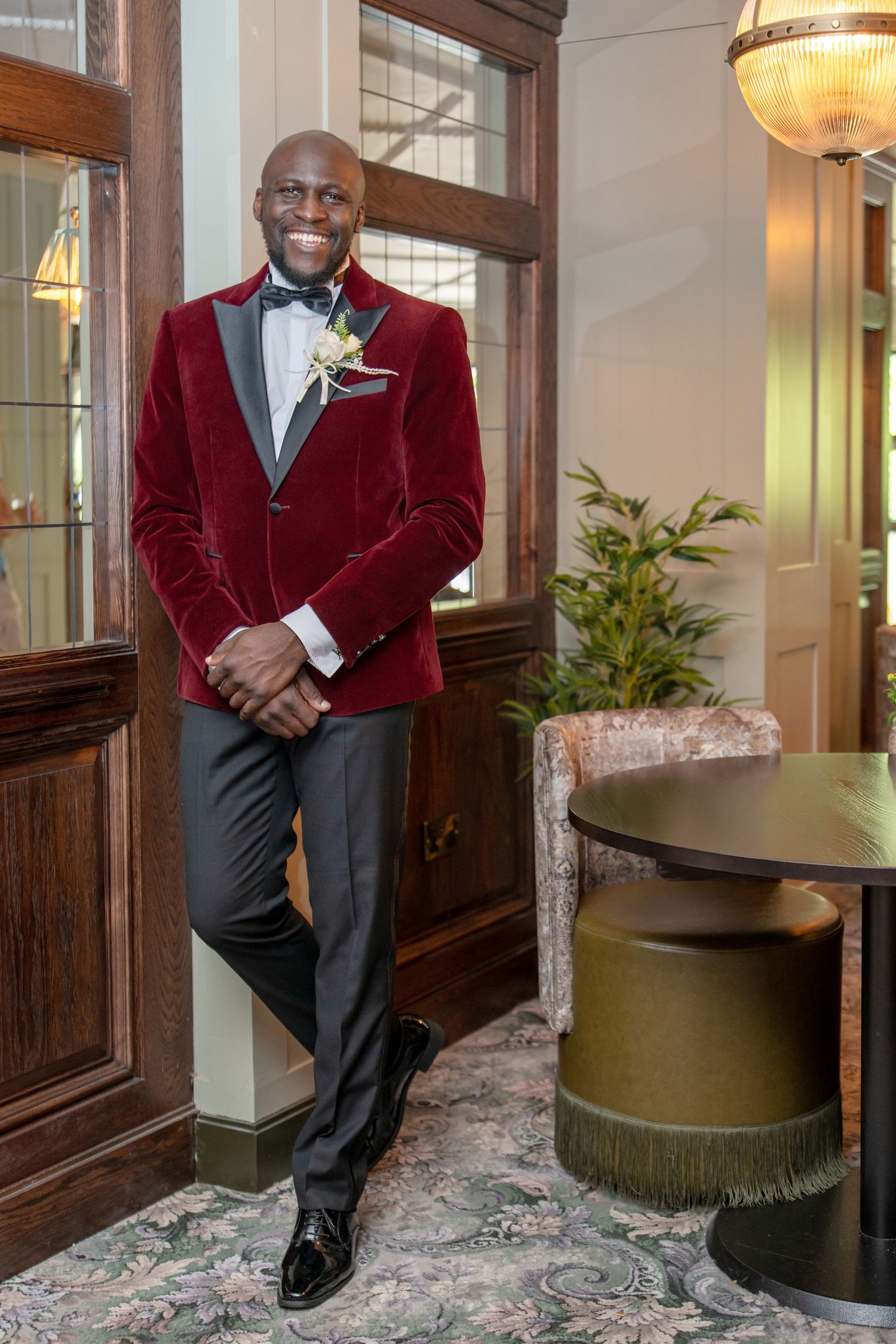 Smiling groom in burgundy velvet tuxedo jacket with boutonniere poses in elegant venue interior.