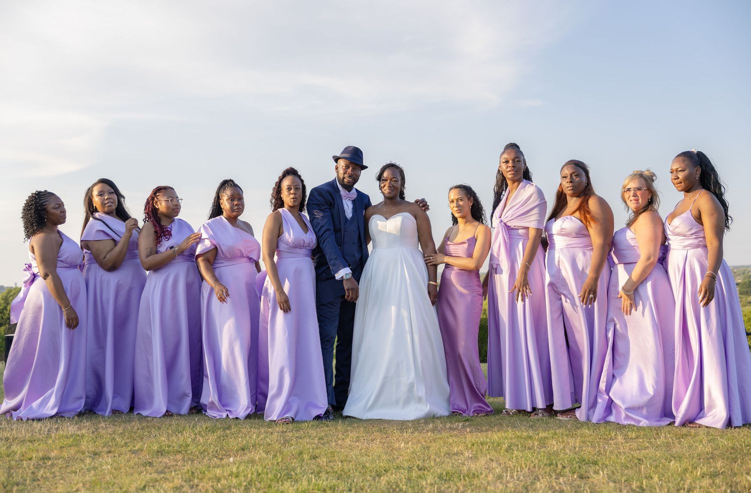 Bride and groom pose with bridesmaids in lavender gowns outdoors on a grassy field under a cloudy sky.
