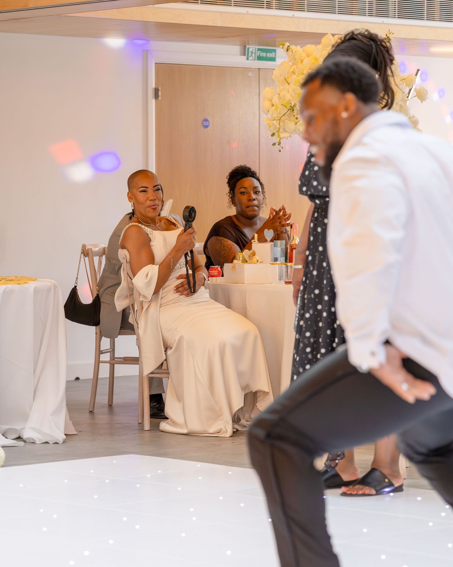 Bride in white dress holds microphone during wedding reception speech while guests watch a dancer perform.