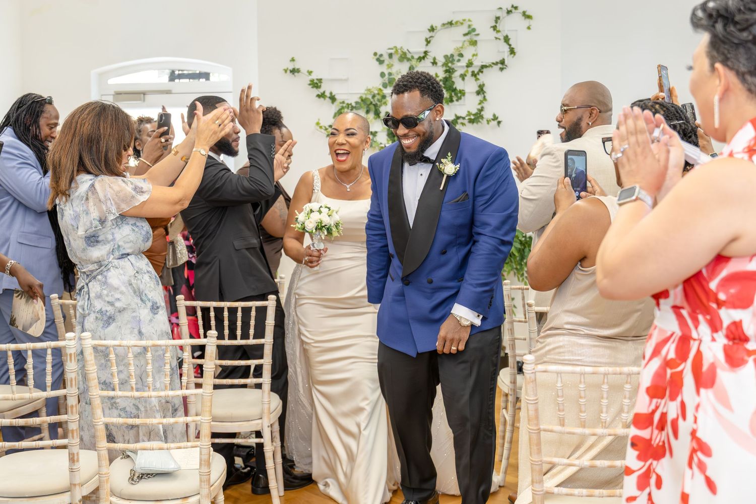 Joyful newlyweds walk down the aisle, groom in blue blazer, bride in white gown, surrounded by celebrating guests.