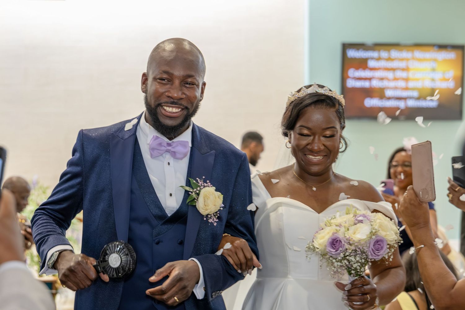 Happy newlyweds walk down the aisle after wedding ceremony, groom in blue suit, bride in white gown holding purple bouquet.