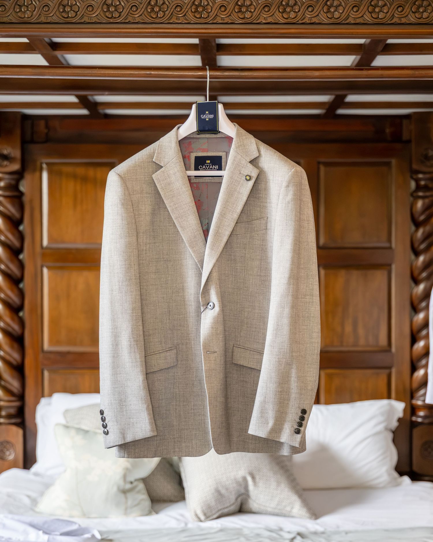 Beige linen blazer hanging on a hanger over an ornate wooden bed with white pillows.