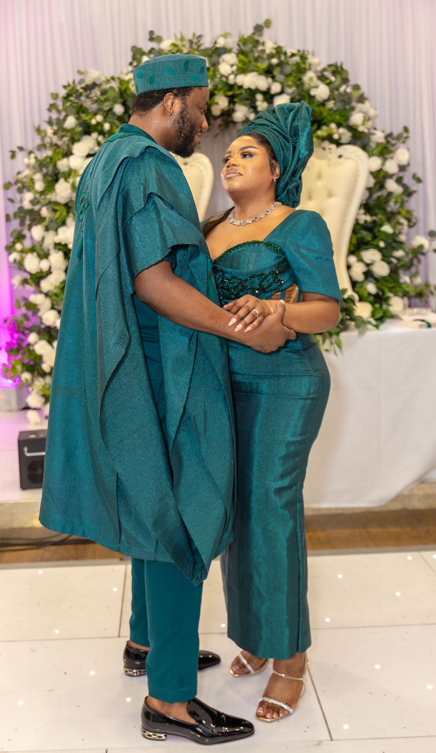 Couple in matching teal traditional African attire embracing near floral arch at wedding celebration.