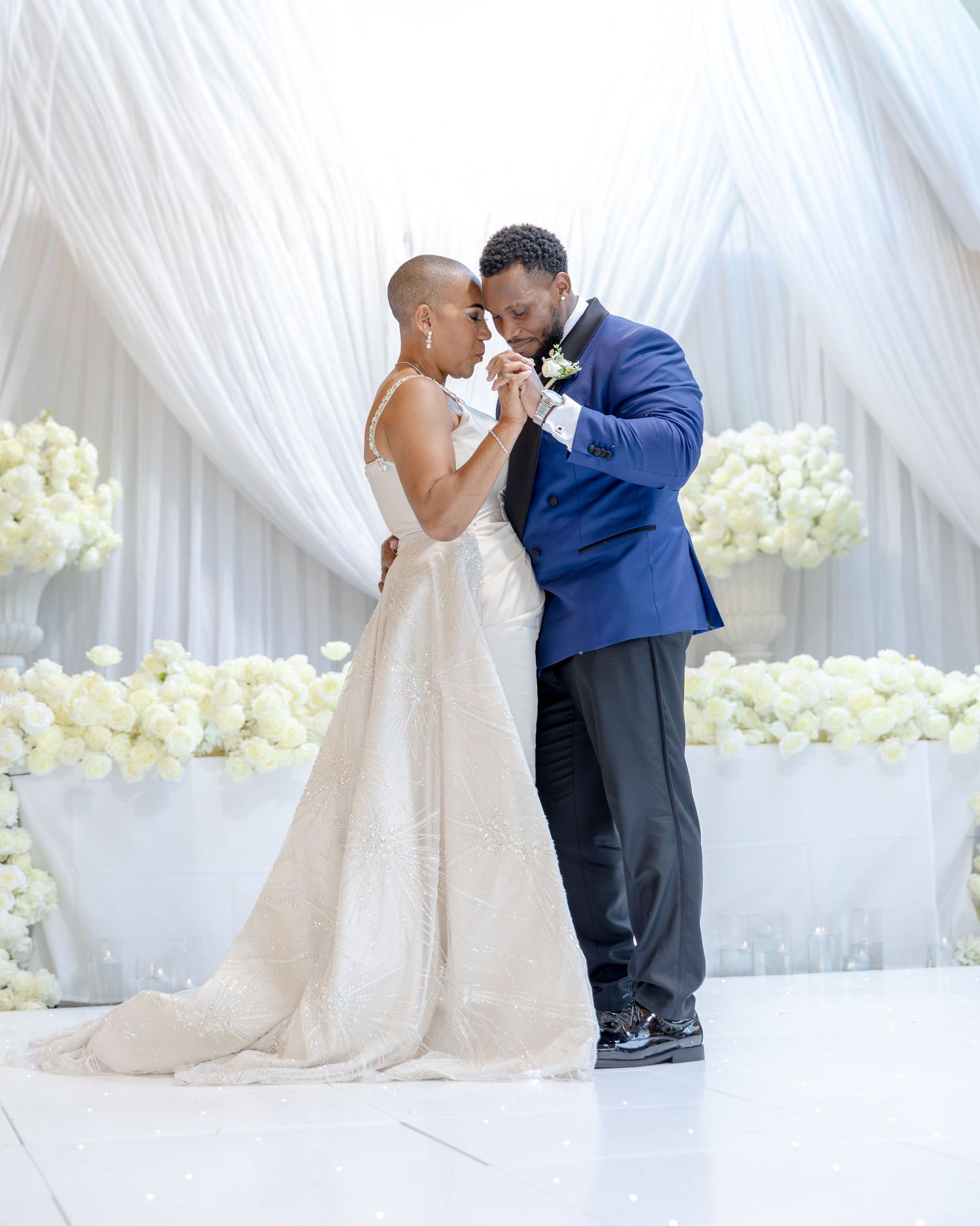 A bride and groom share their first dance at an elegant wedding reception with white floral arrangements and draped fabric backdrop.