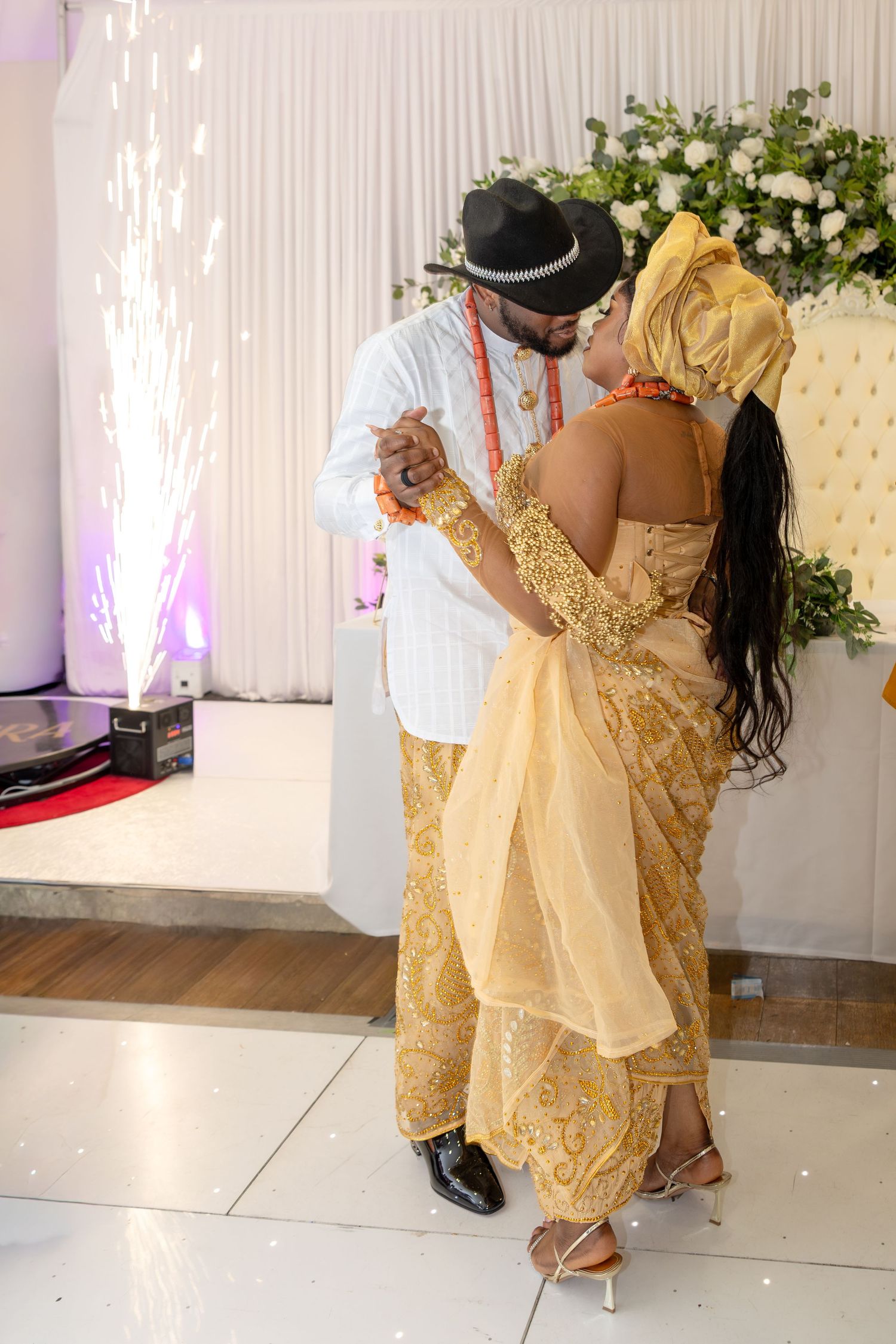 Nigerian couple sharing first dance in traditional wedding attire with gold aso-oke and white agbada amid sparkling fireworks.