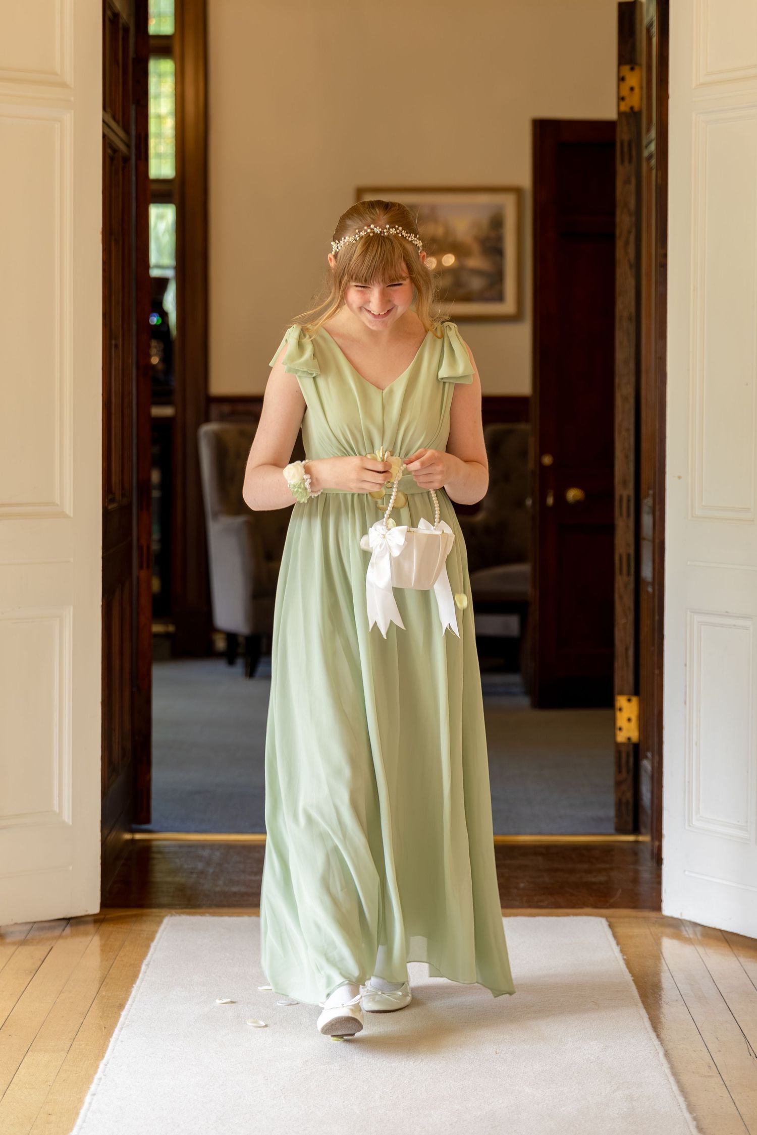 Young woman in sage green bridesmaid dress with floral crown holding white ribbon basket walks through elegant doorway.
