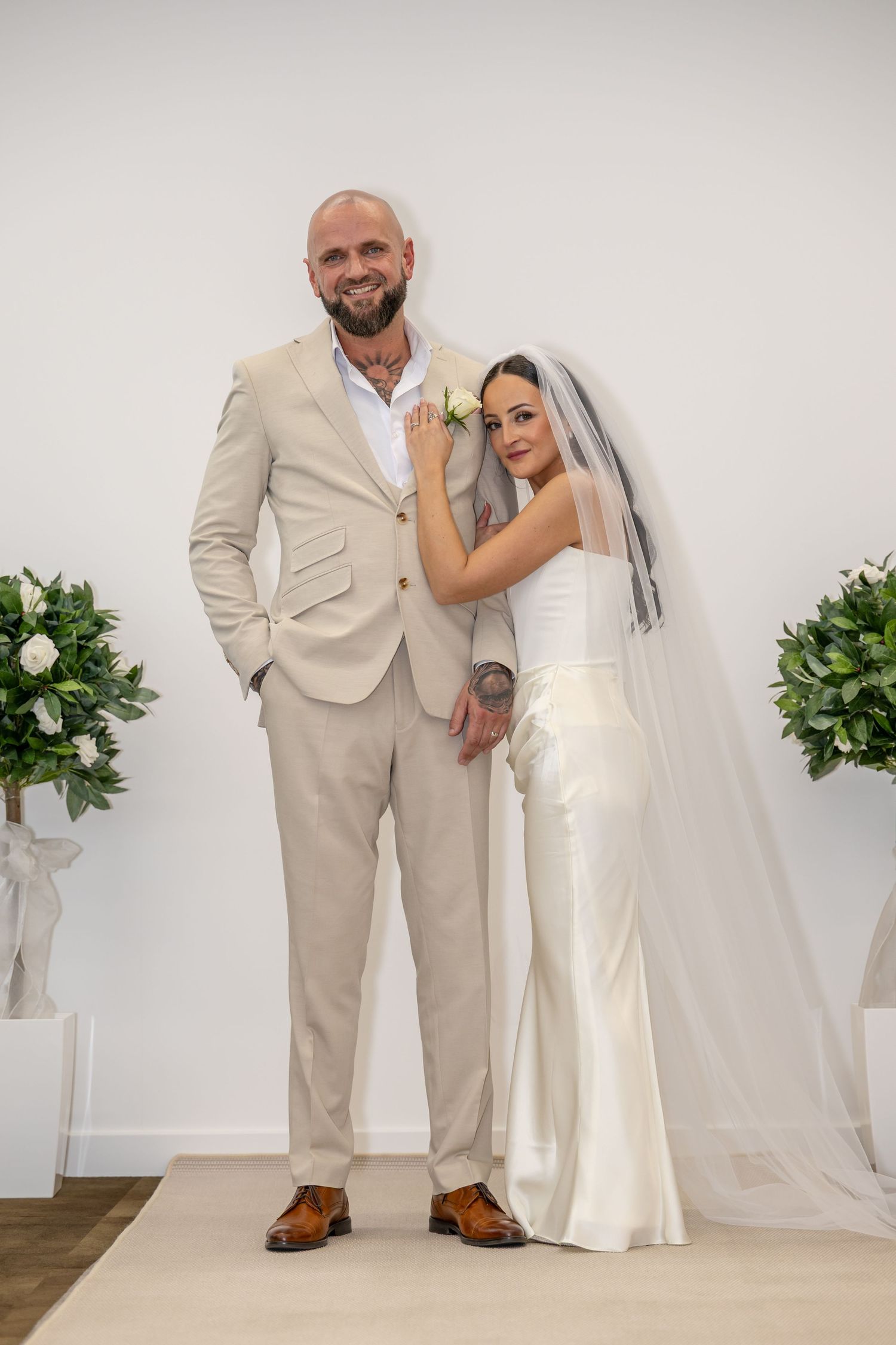Bride in white satin gown and veil standing with groom in beige suit at wedding altar.
