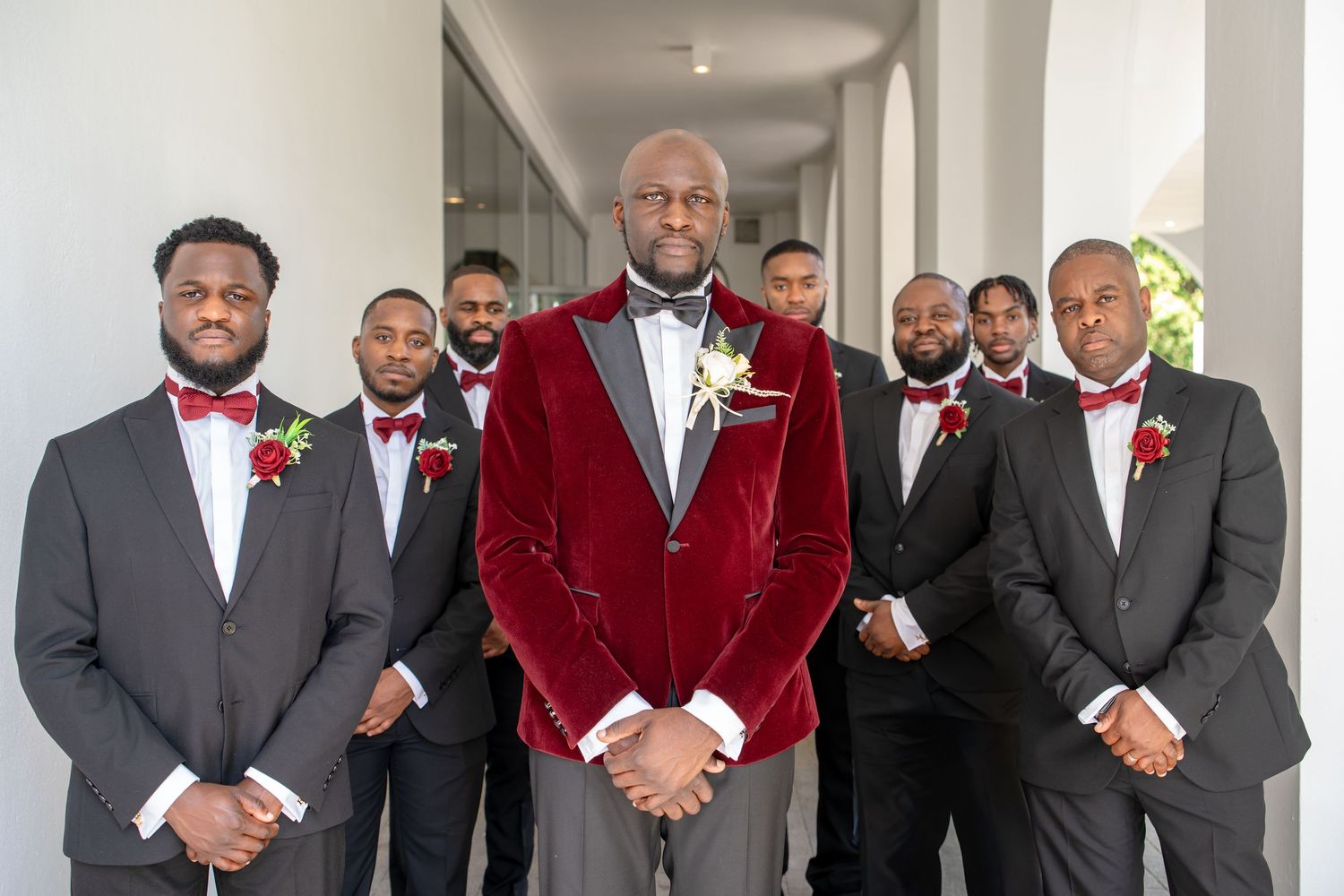 Groom in red velvet tuxedo jacket poses with groomsmen in black suits and red bow ties at wedding.