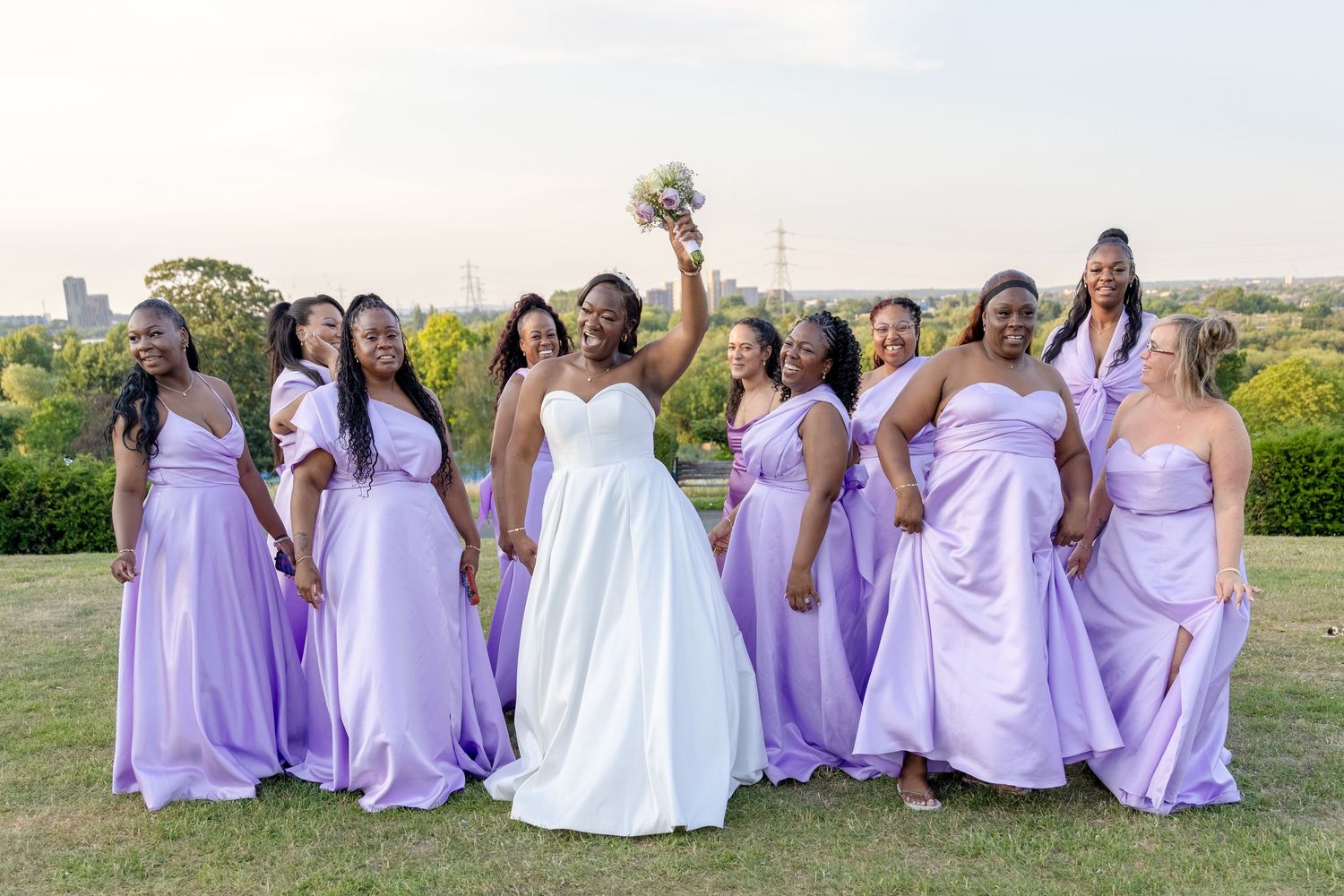 Bride in white gown celebrates with bridesmaids in lavender dresses outdoors, bouquet raised joyfully.