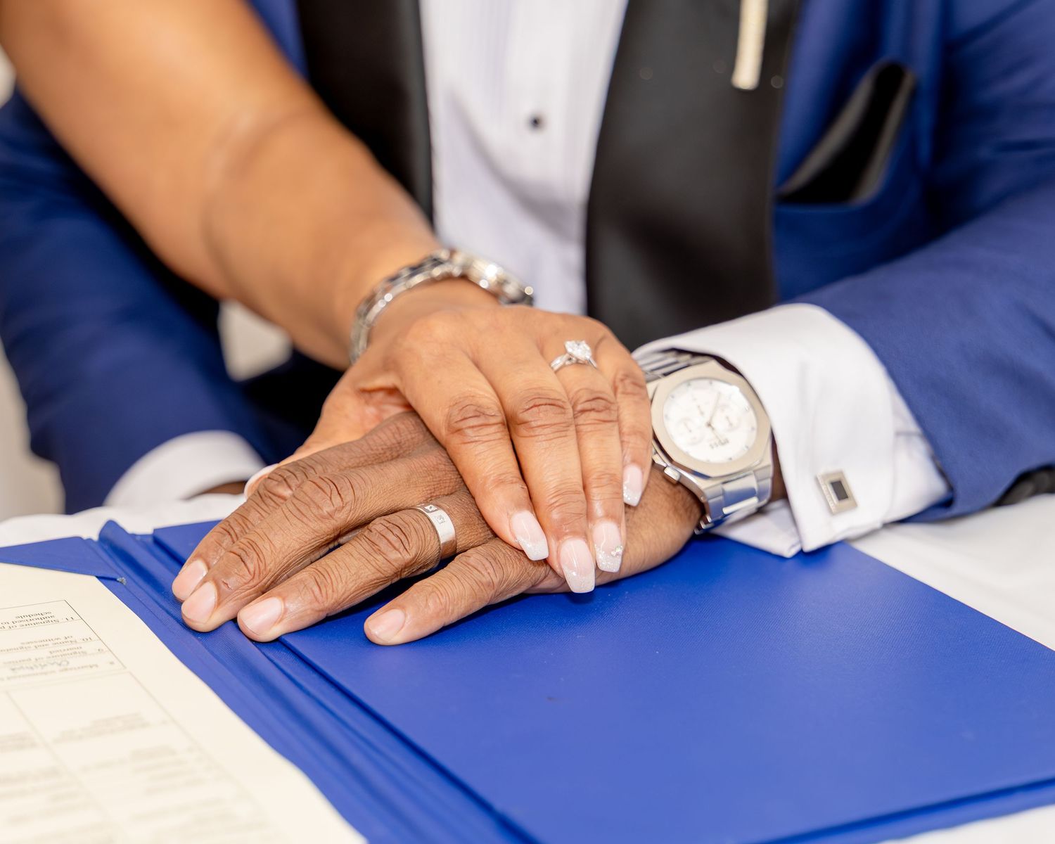Couple placing hands together showing wedding rings and watch on blue folder at signing ceremony.