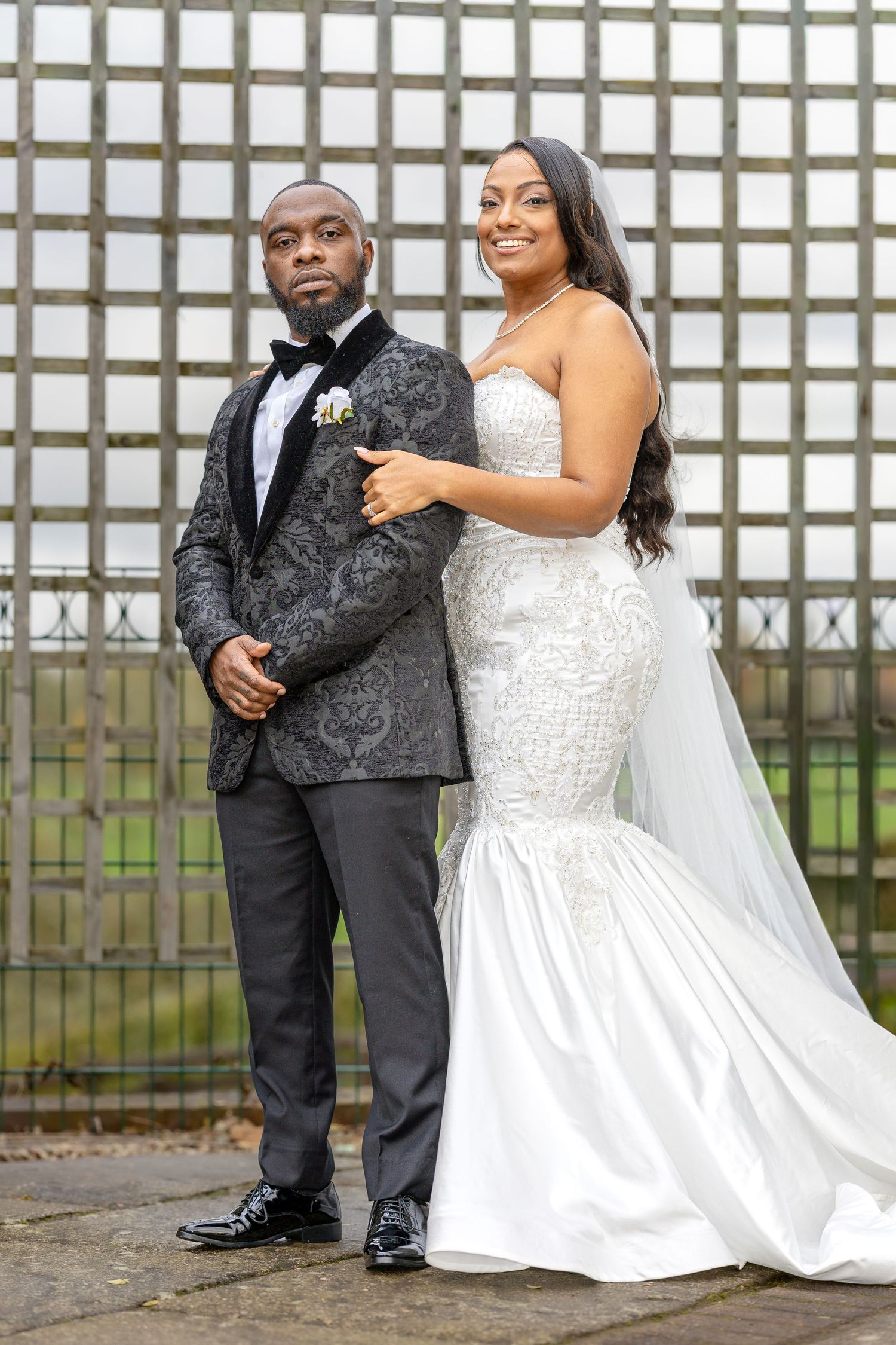 A newlywed couple poses together, with the bride in a white mermaid gown and groom in a patterned tuxedo jacket.