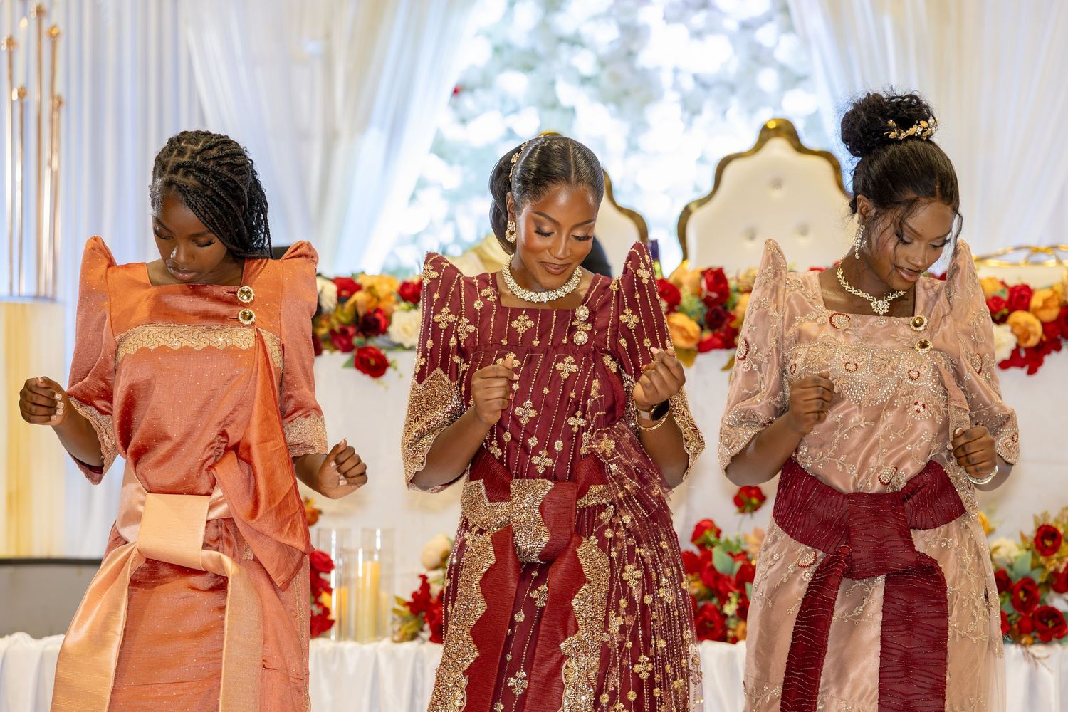 Three young women in matching orange and red African traditional attire bow their heads in prayer at ceremony.