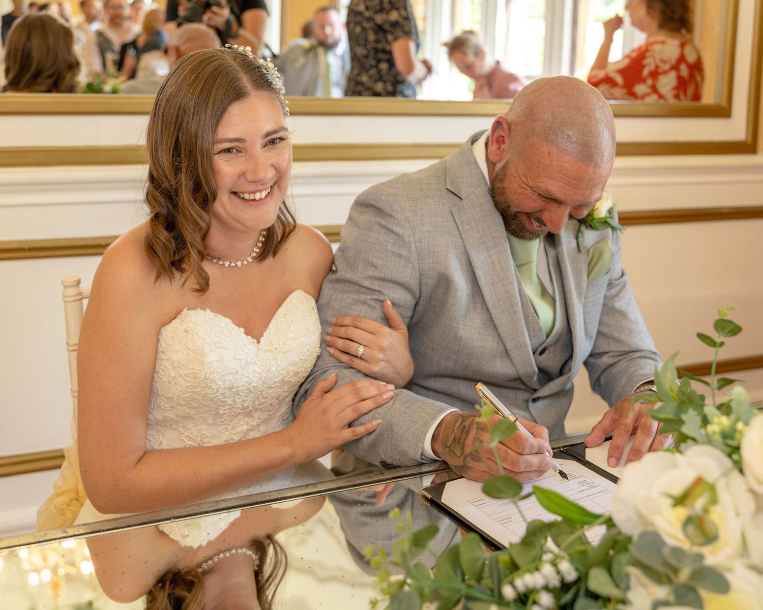 Bride smiling joyfully as groom signs wedding register at elegant reception venue.