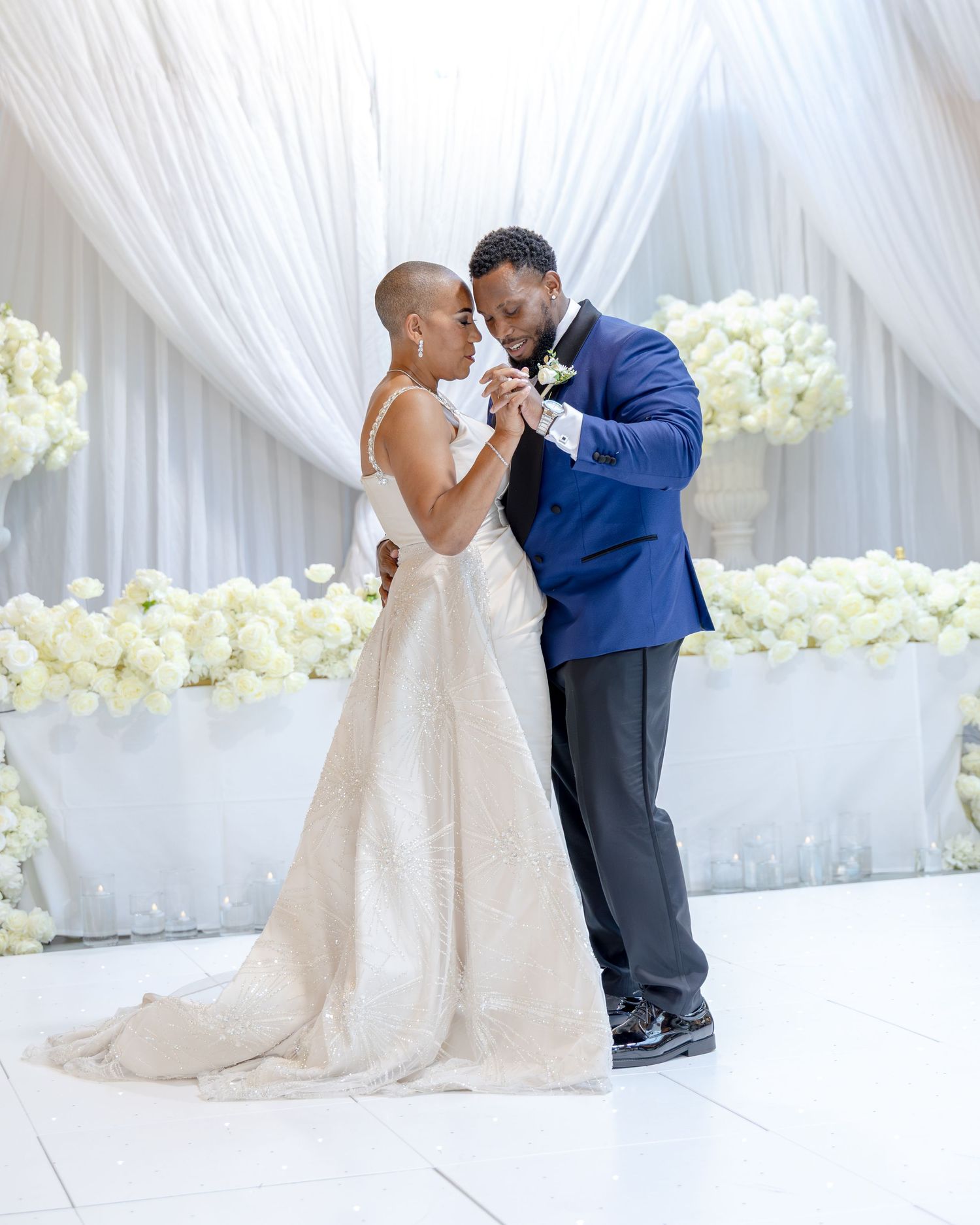 Bride and groom share first dance at elegant wedding reception with white floral arrangements and draped backdrop.