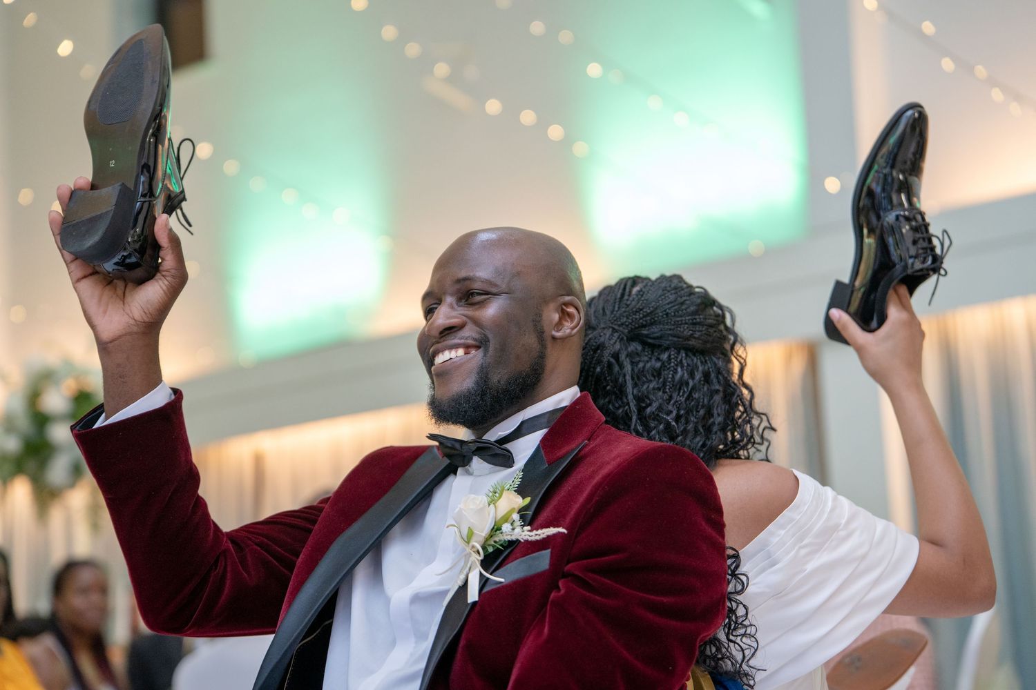 Smiling groom in red velvet tuxedo holds up shoes during wedding reception shoe game with bride.