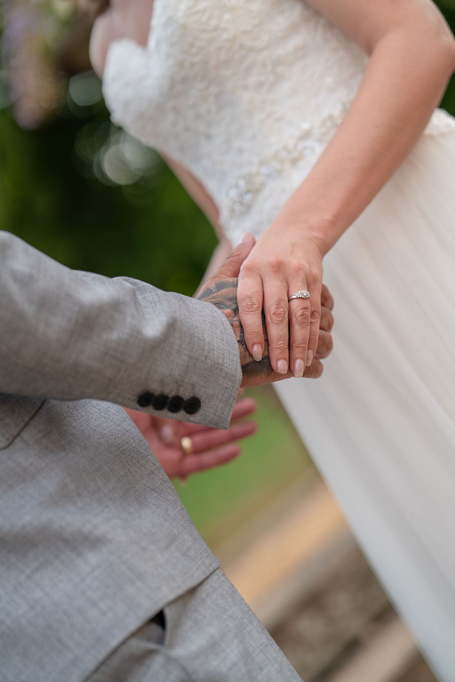 Bride and groom holding hands at wedding ceremony, showing diamond engagement ring and wedding bands, groom in gray suit.