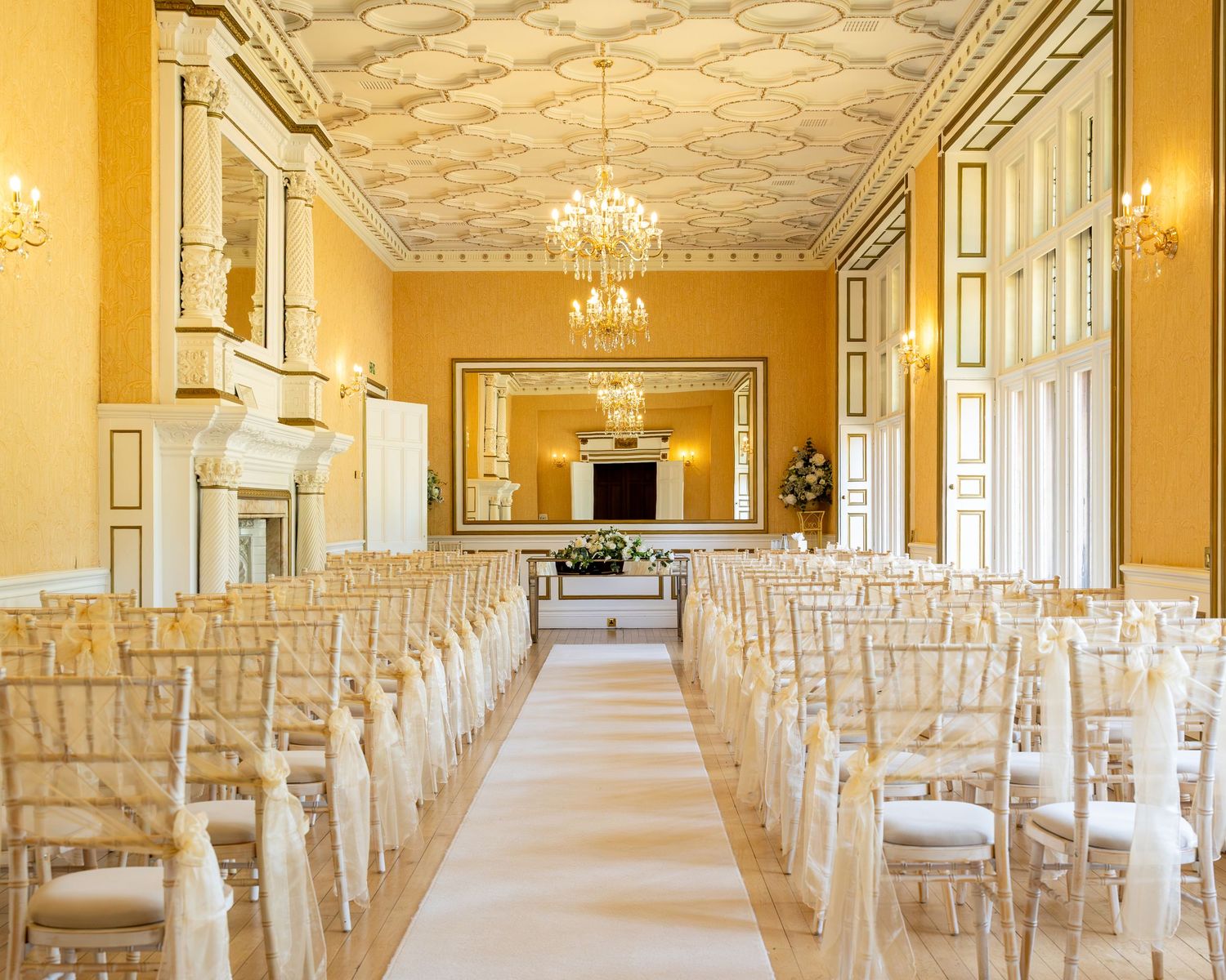Elegant wedding ceremony hall with gold walls, ornate ceiling, chandelier, and rows of chiavari chairs lining a white aisle runner.
