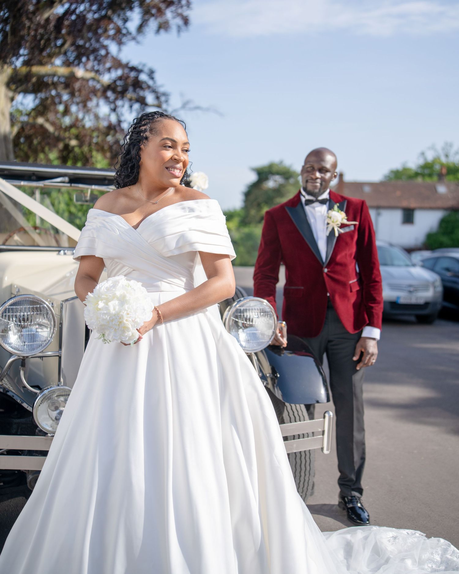 Bride in white off-shoulder gown holds bouquet as groom in red velvet blazer approaches vintage car outdoors.