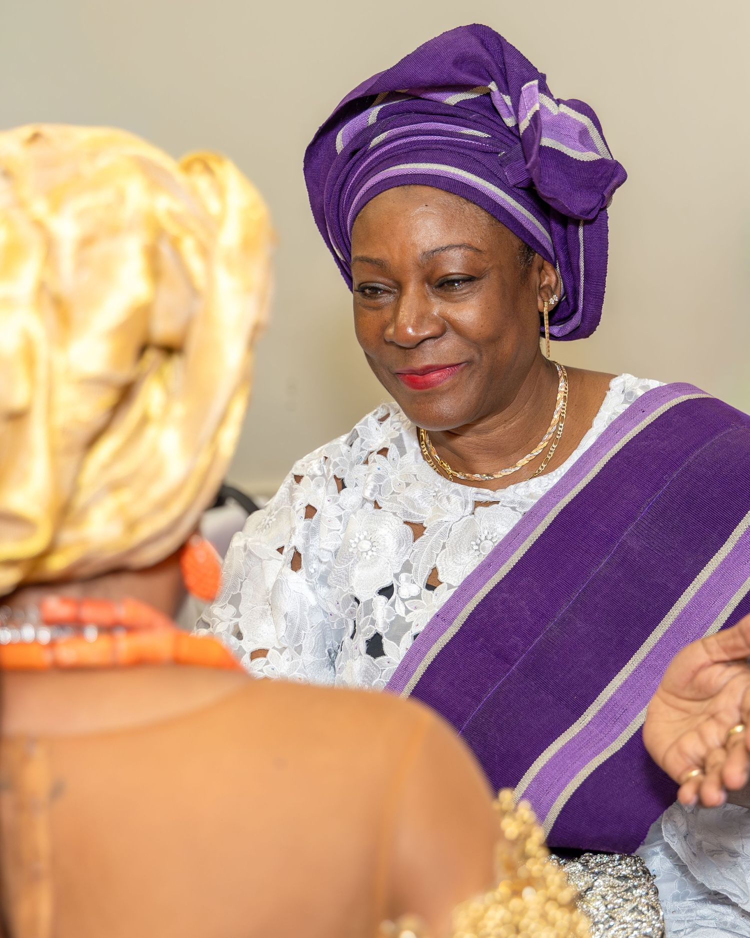 Elegant Nigerian woman in purple gele and white lace smiles warmly at bride during traditional ceremony.