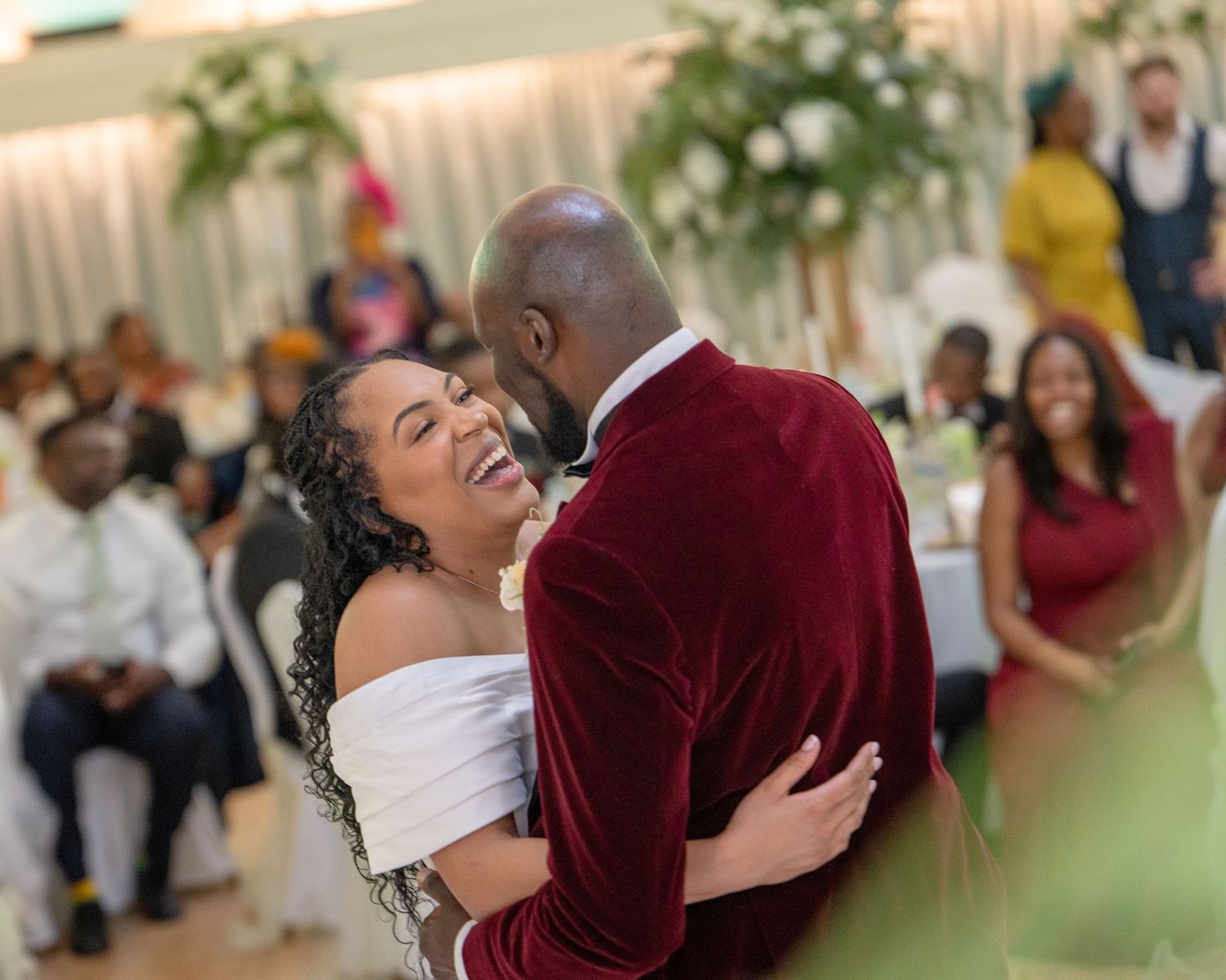 Joyful couple sharing a first dance at a wedding reception, bride in white off-shoulder gown laughing with groom in burgundy velvet suit.