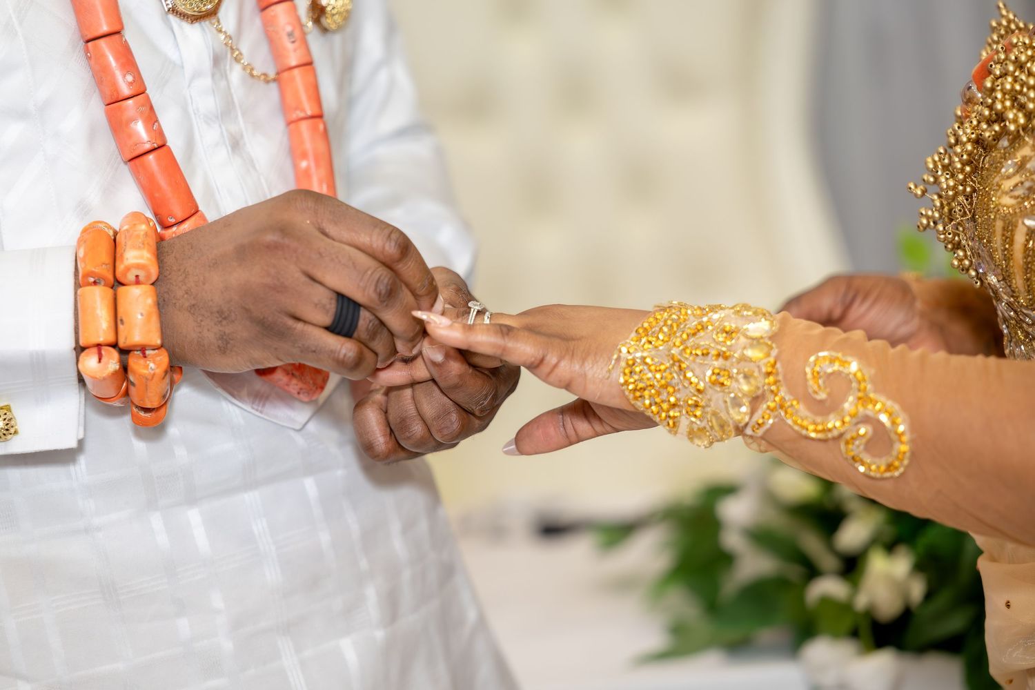 Close-up of Nigerian traditional wedding ring exchange, groom wearing coral beads and white attire, bride with gold henna hands.