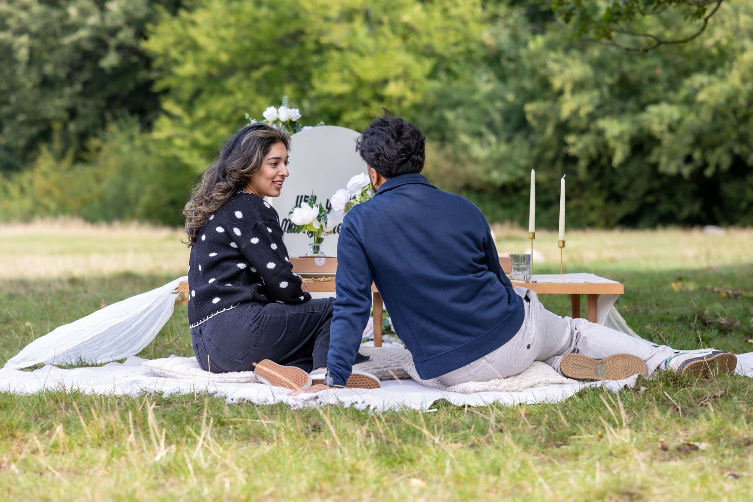 Couple sitting together on a white picnic blanket in a scenic green park during a romantic proposal.
