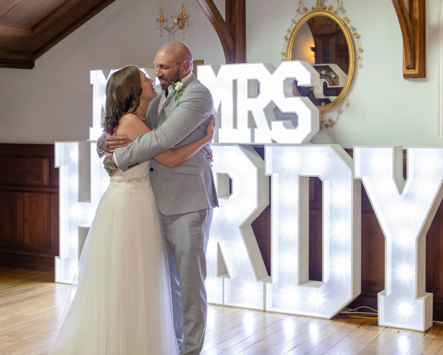 Newlyweds embrace in front of illuminated MRS HARDY marquee letters at elegant wedding reception venue.