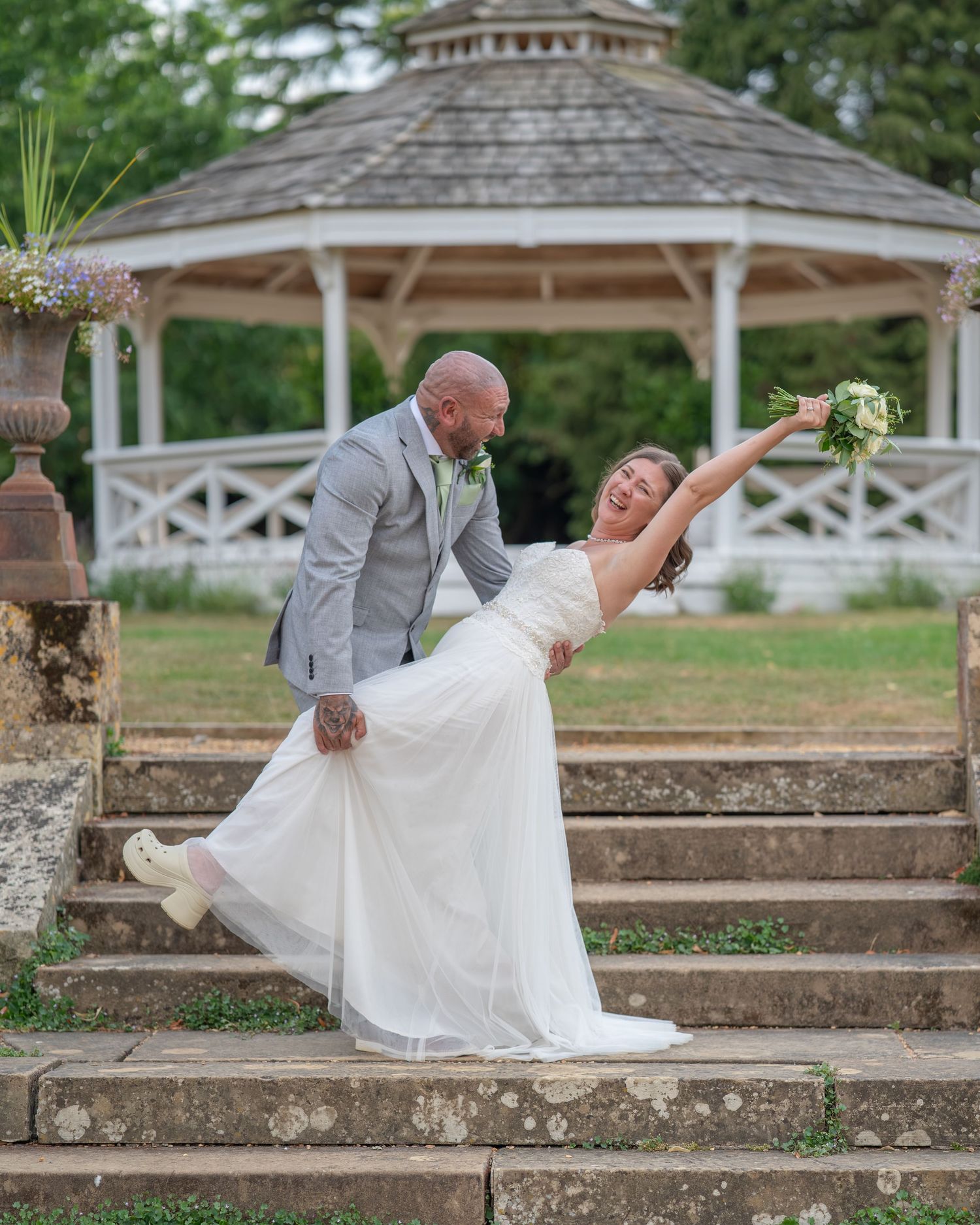 Groom dips bride on stone steps before a white gazebo in a lush garden during an outdoor wedding ceremony.