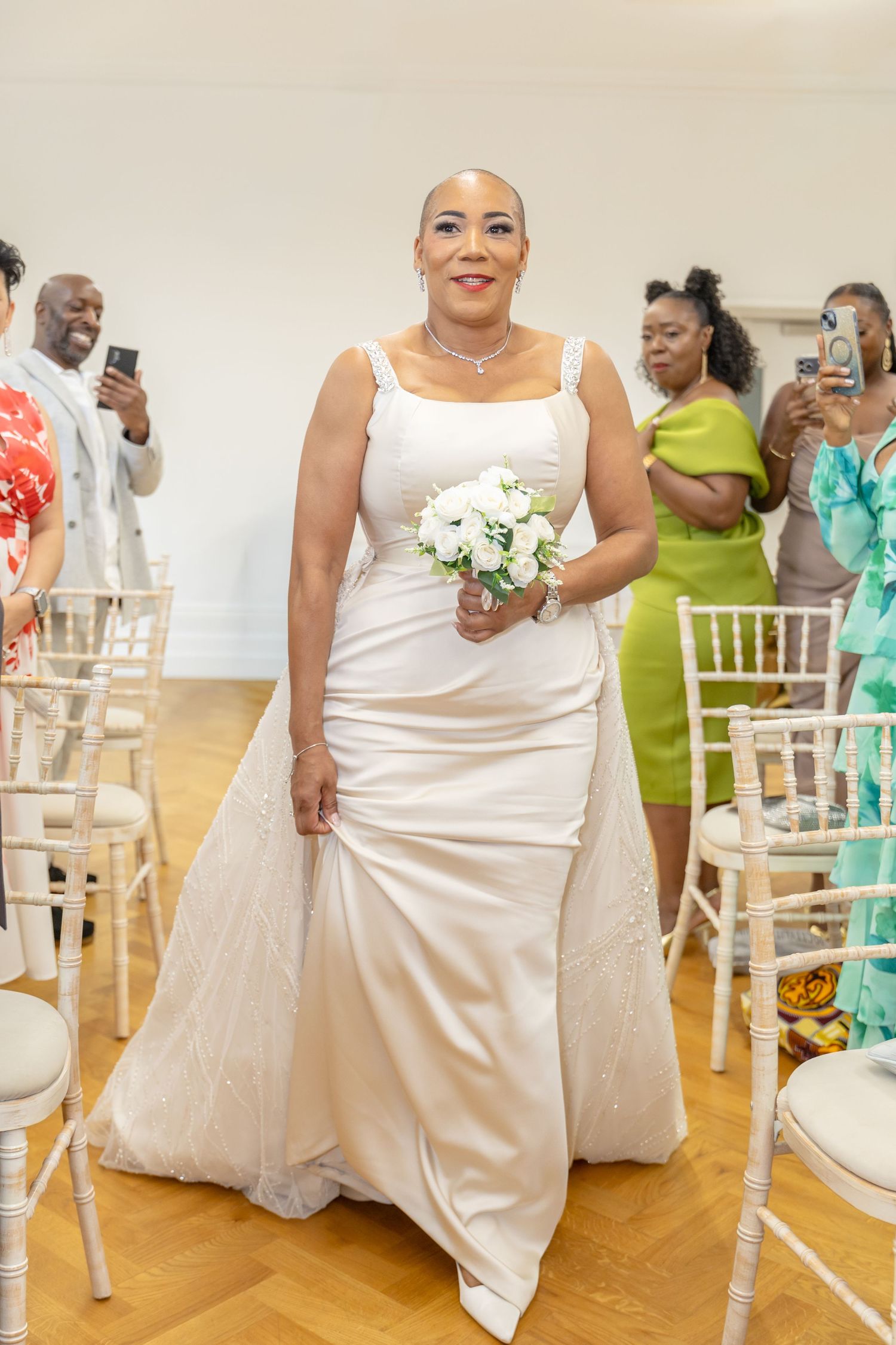 Bride in elegant white satin gown holding bouquet walks down aisle at wedding ceremony with guests photographing.