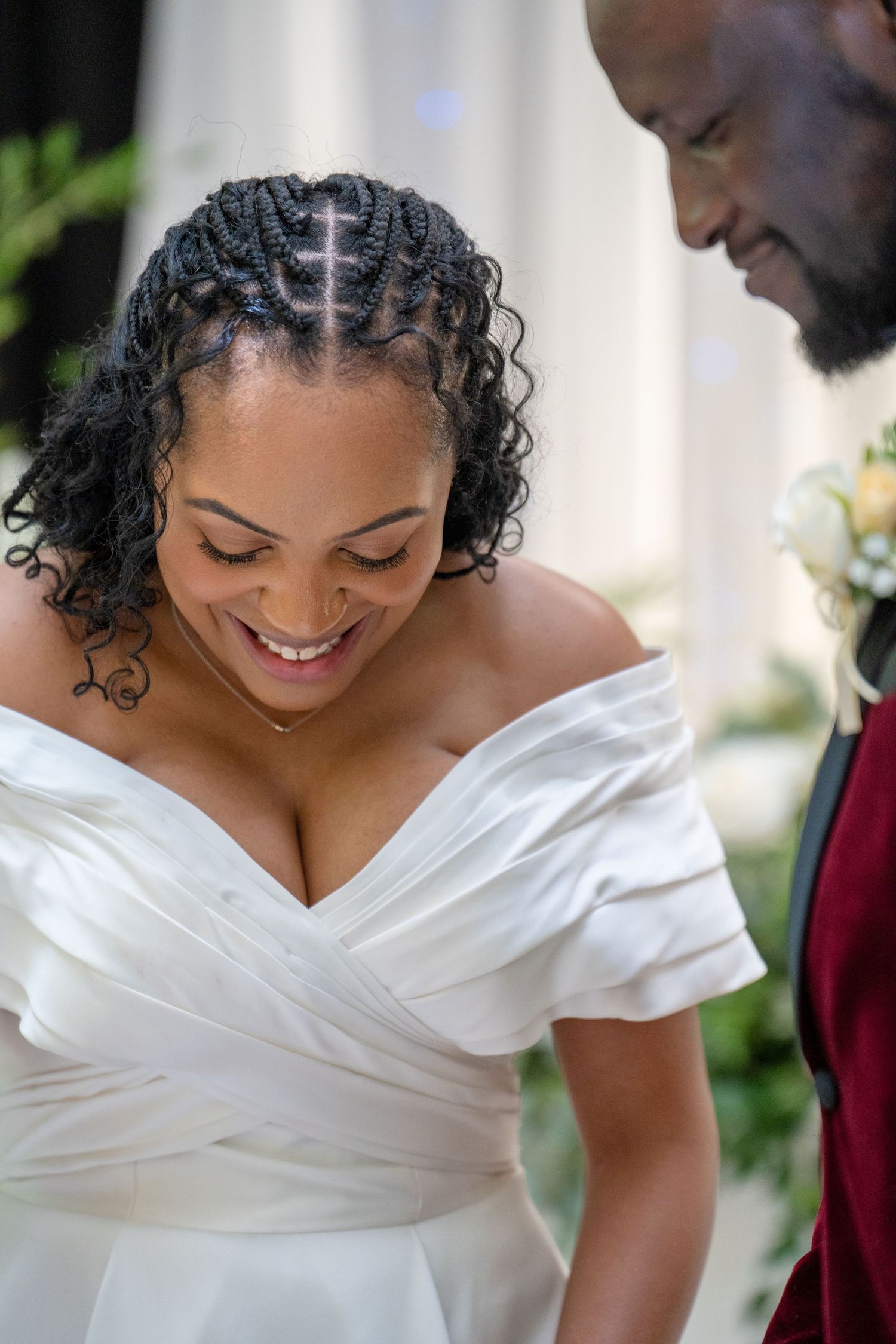 Smiling Black bride in white off-shoulder gown stands beside groom in burgundy suit during wedding ceremony.