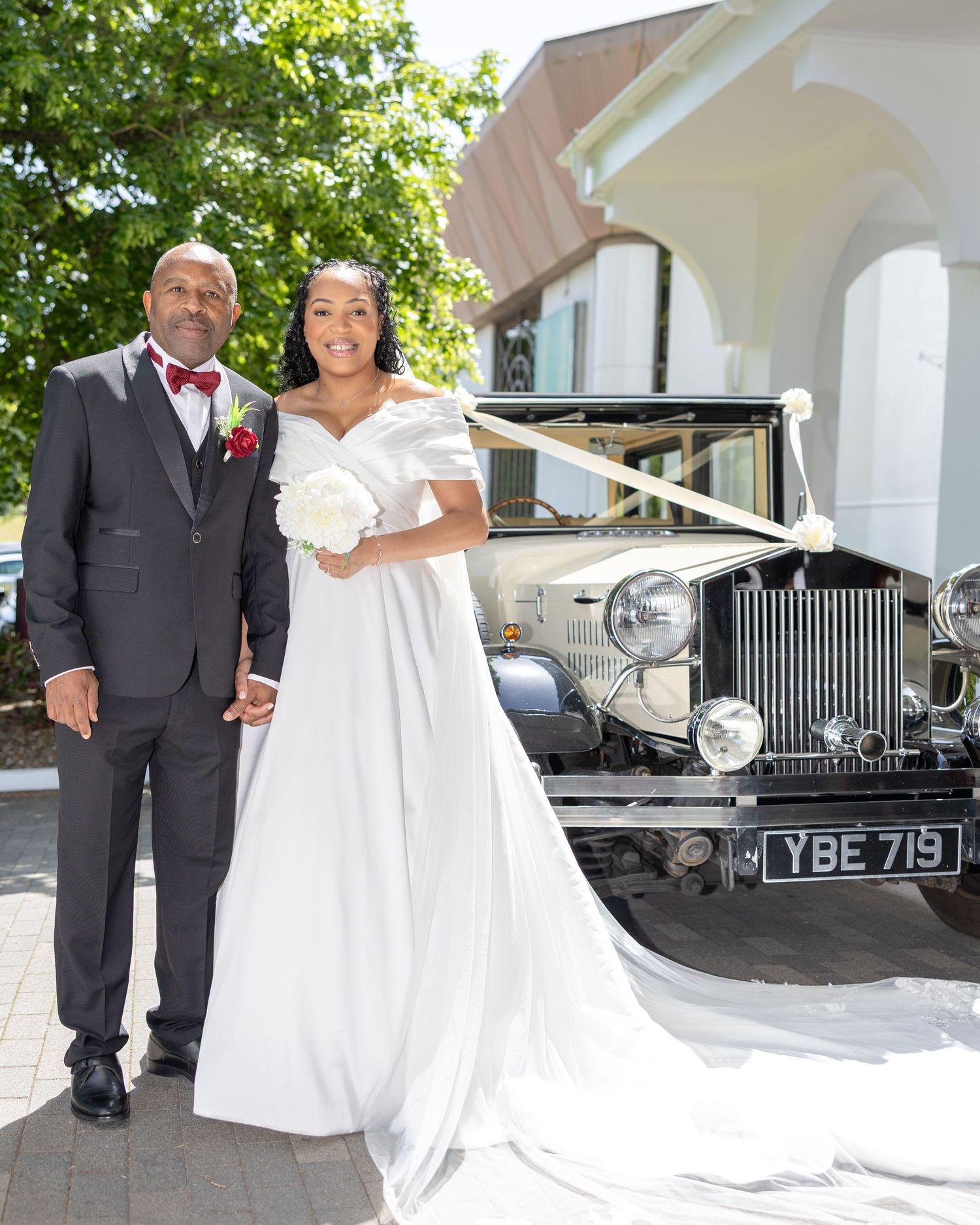 Bride and groom posing beside a vintage Rolls-Royce decorated with white ribbons outside a church.