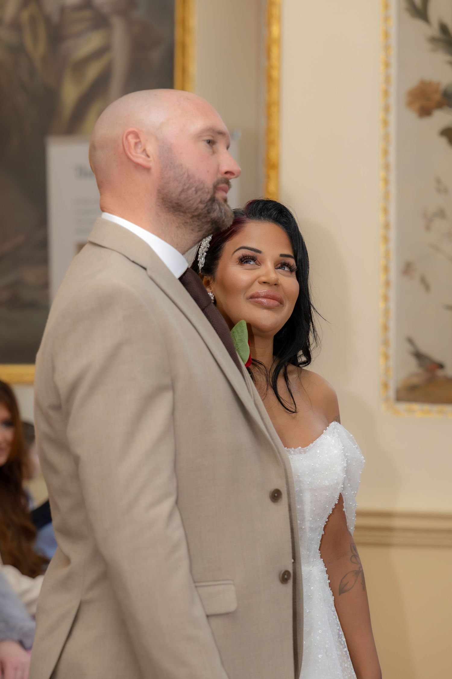 Bride and groom standing together during wedding ceremony, bride smiling warmly in white dress, groom in beige suit.