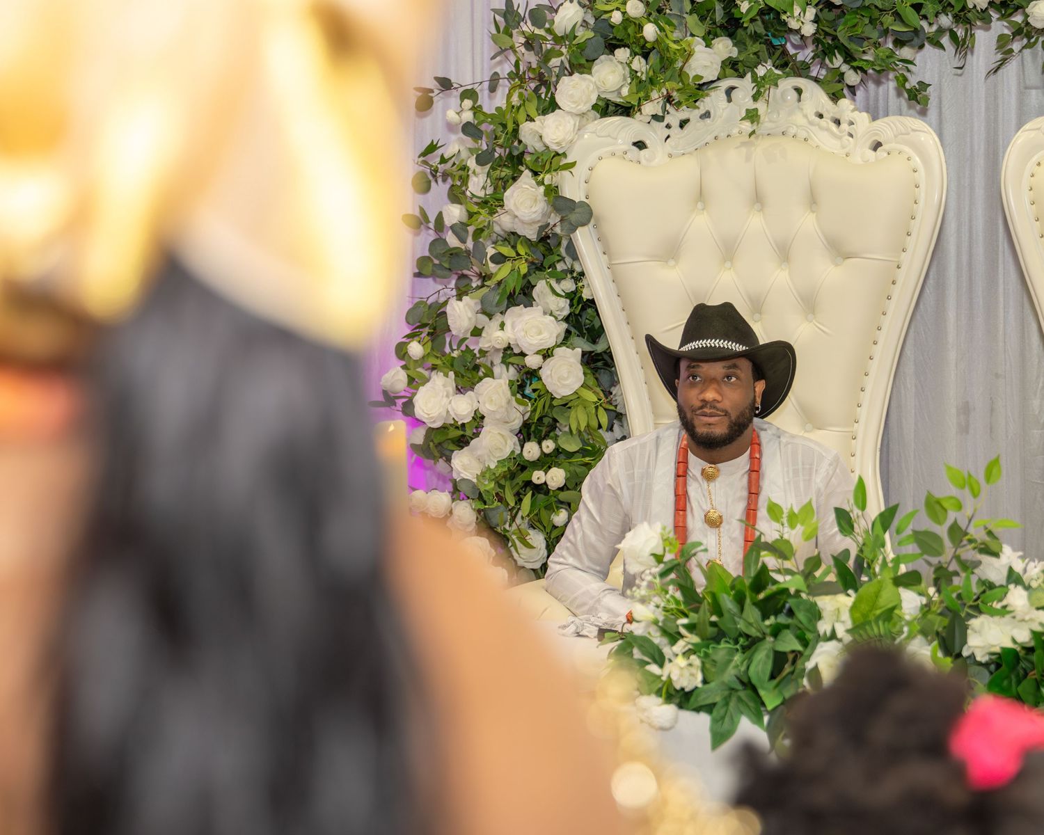 Groom in black cowboy hat and gold beads sits on ornate white throne chair at Nigerian traditional wedding.