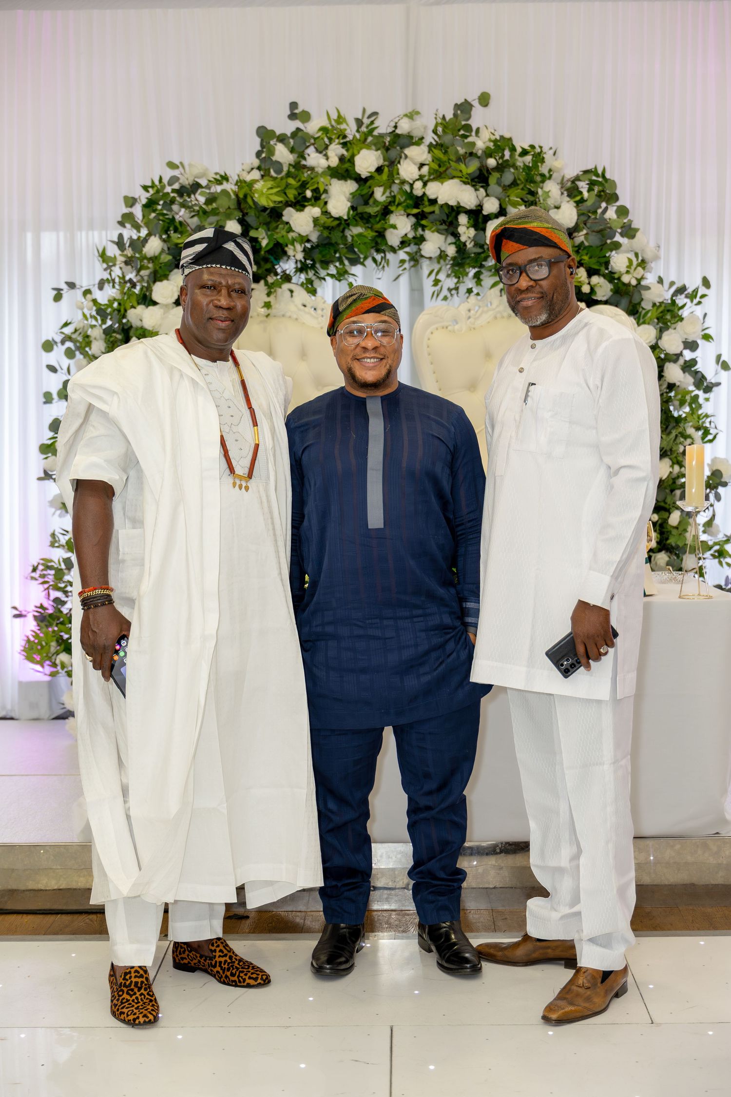 Three men in Nigerian traditional attire posing before a white floral arch at a wedding reception.