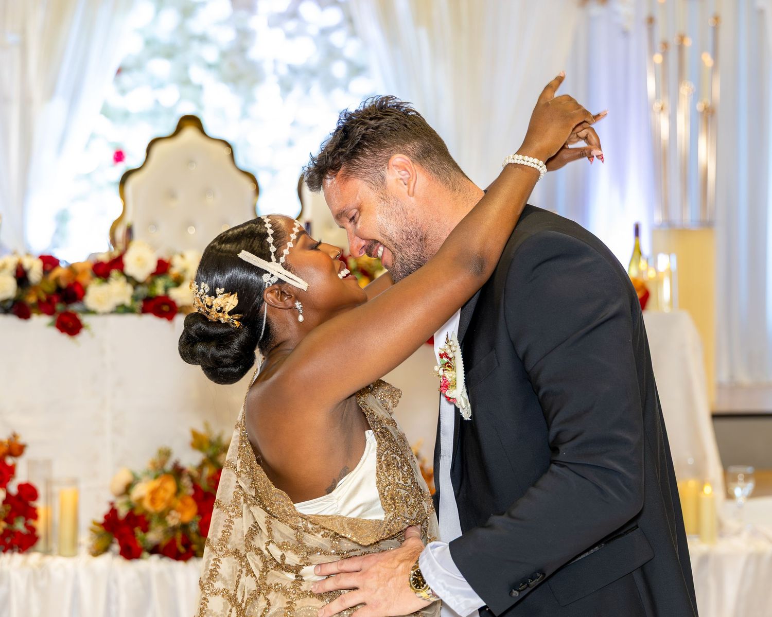 Interracial couple shares romantic first kiss at elegant wedding reception with floral backdrop.