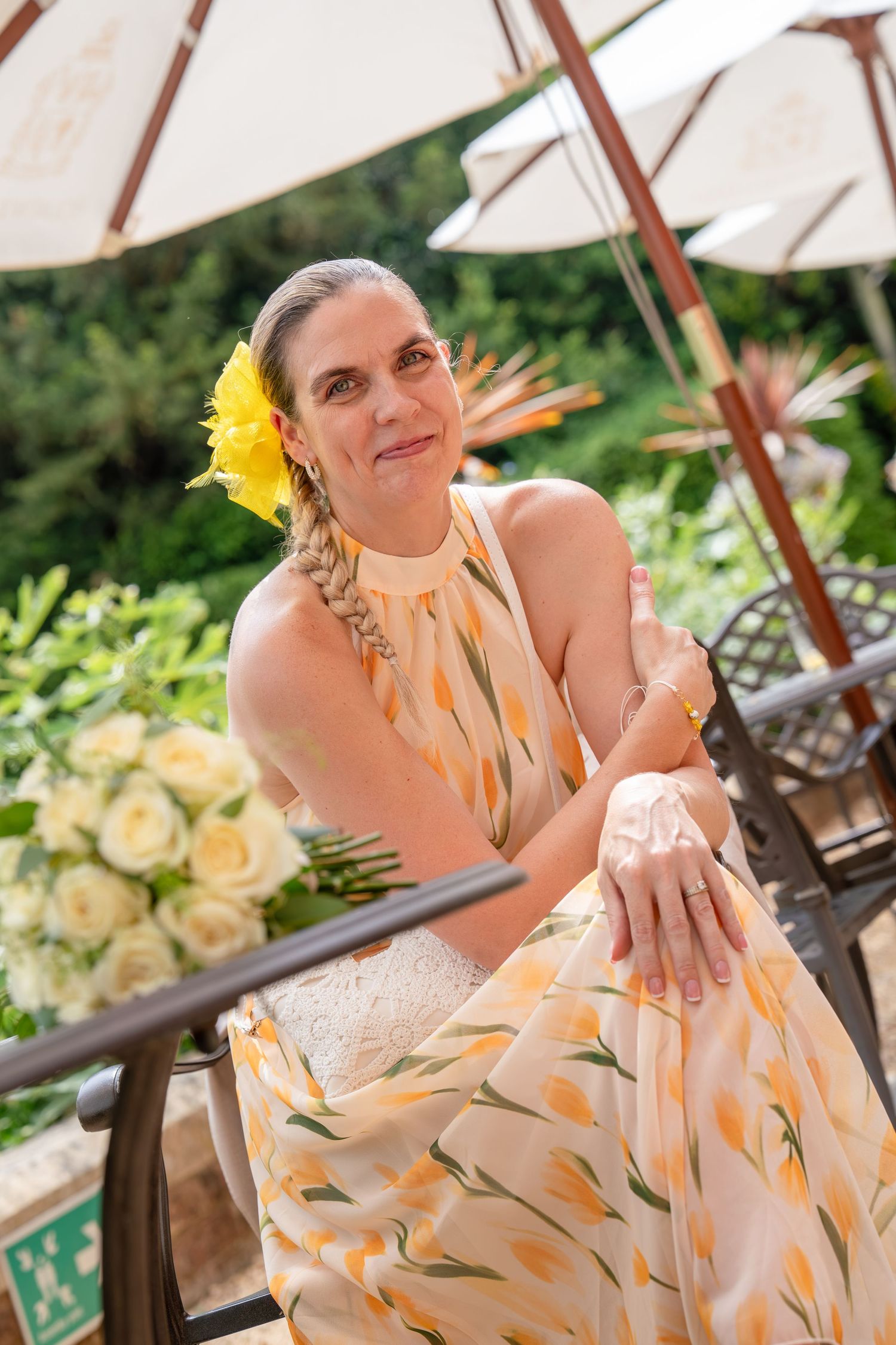 Woman in yellow floral dress with flower in hair sits outdoors near white roses and patio umbrellas.