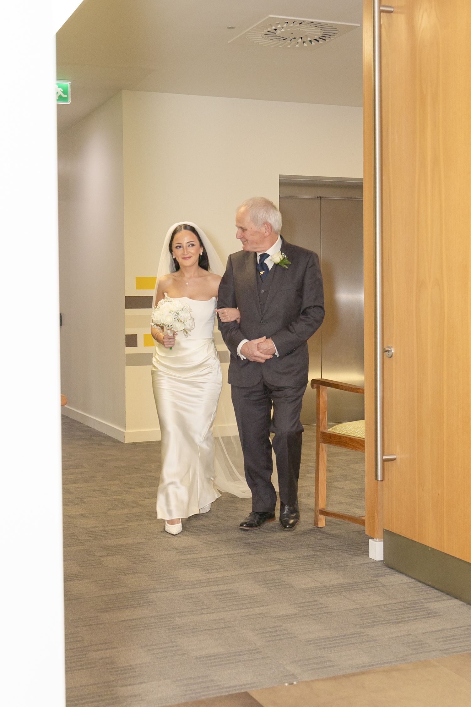 Bride in white gown walks arm-in-arm with older man in grey suit through modern venue corridor.