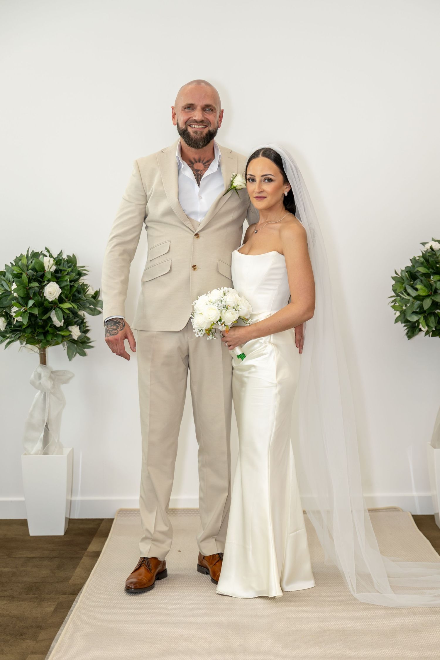 Bride in white satin gown and groom in beige suit pose together in bright minimalist venue.