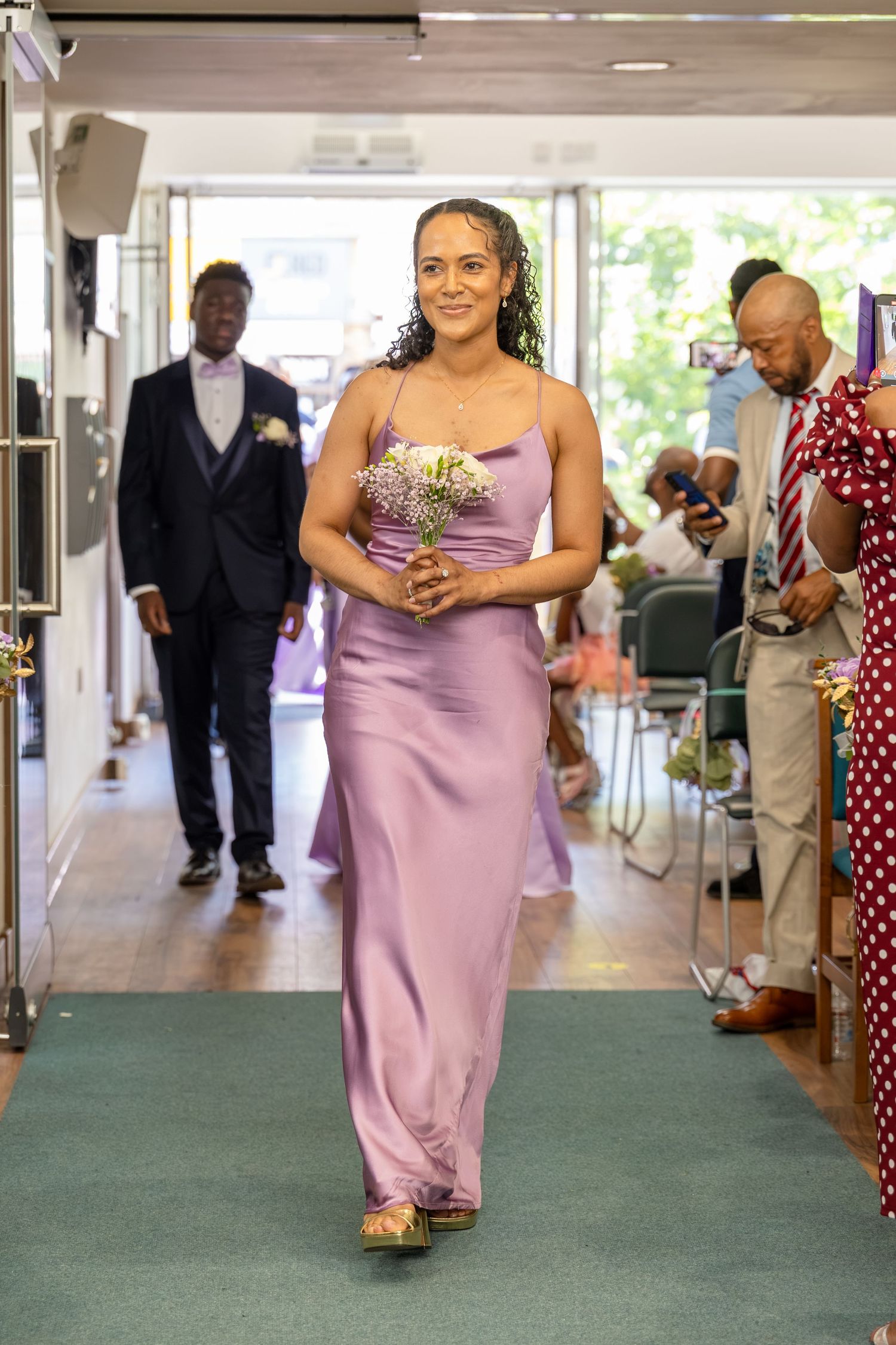 Bridesmaid in lavender satin gown walks down aisle holding bouquet at wedding ceremony.