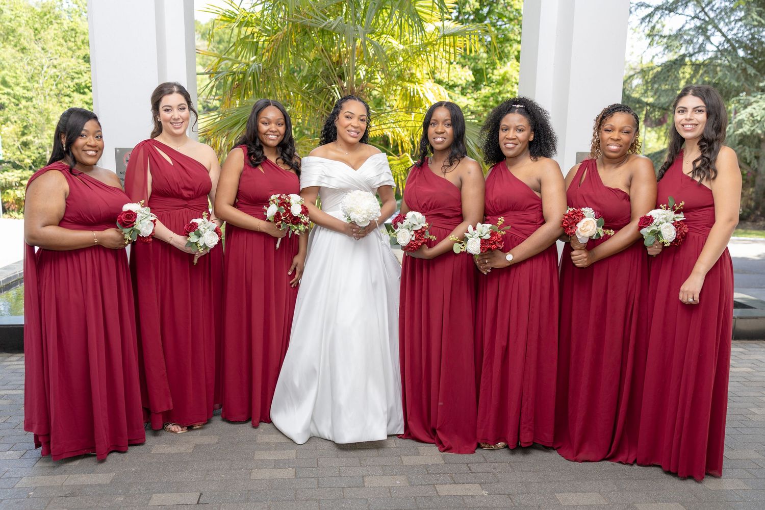 Bride in white gown poses with eight bridesmaids in matching red dresses holding white bouquets outdoors.