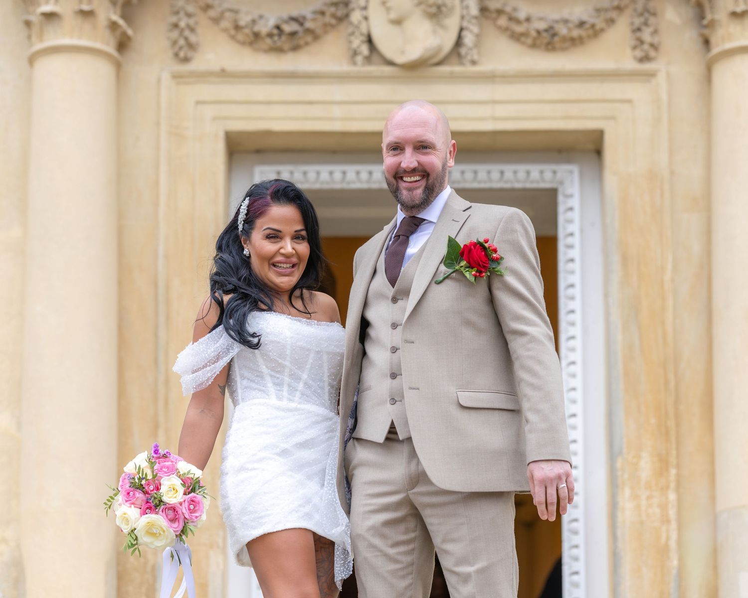 Bride and groom pose at ornate stone doorway, bride holding pink bouquet in white mini dress.