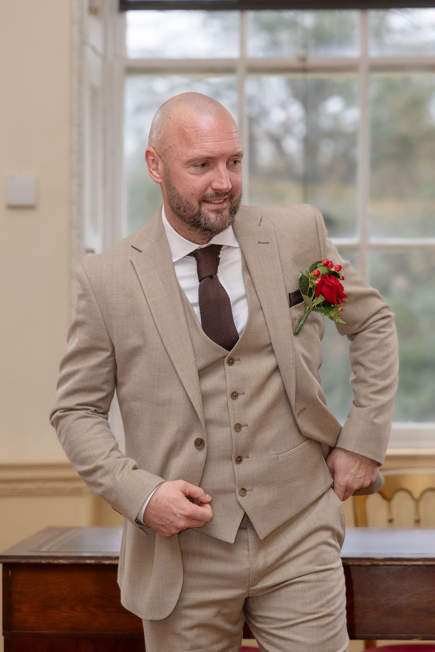 Bald groom in elegant beige three-piece suit with red boutonniere smiling at a wedding ceremony.