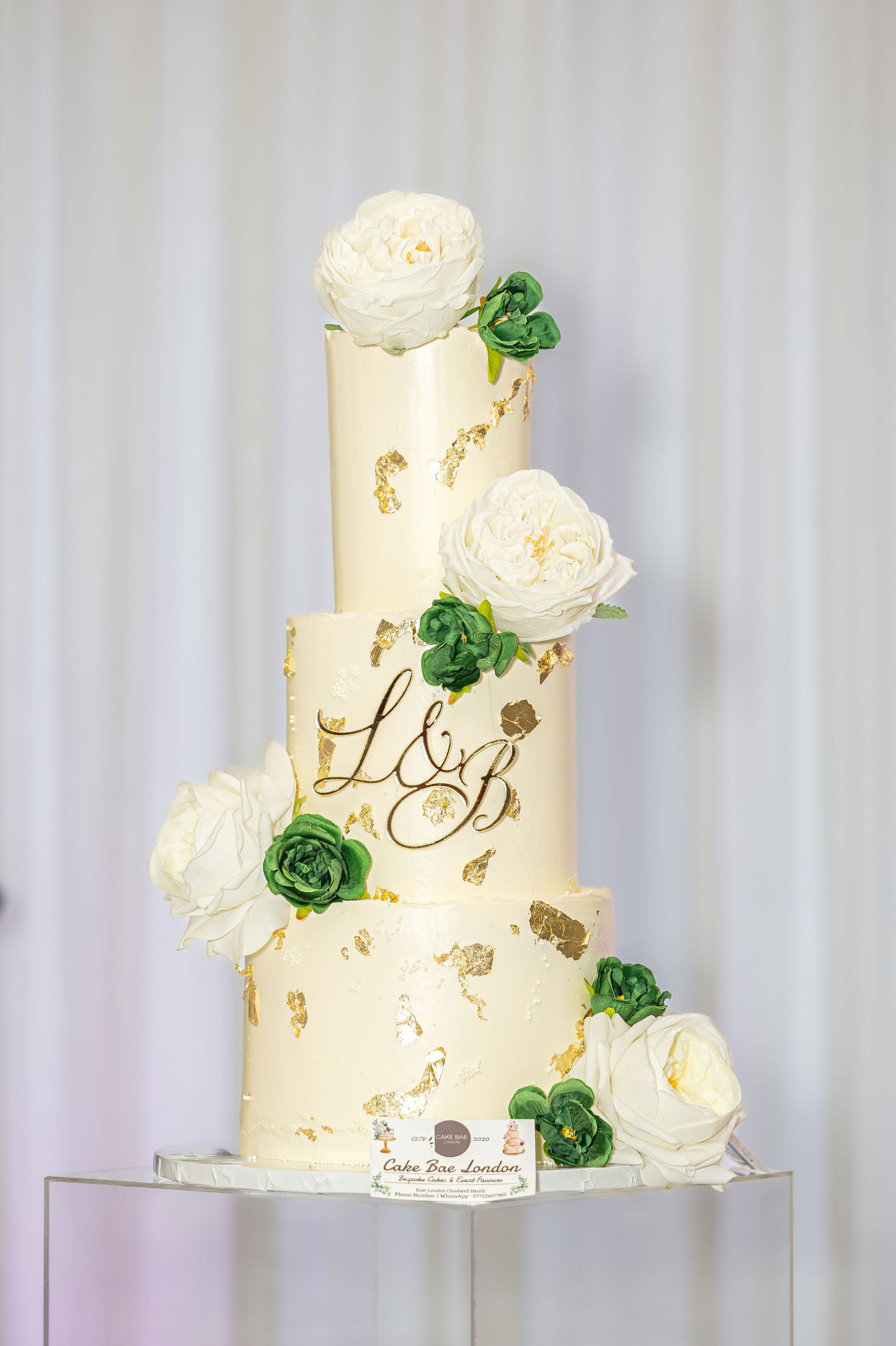 Three-tier ivory wedding cake with gold leaf, monogram L&B, and white and green floral decorations.