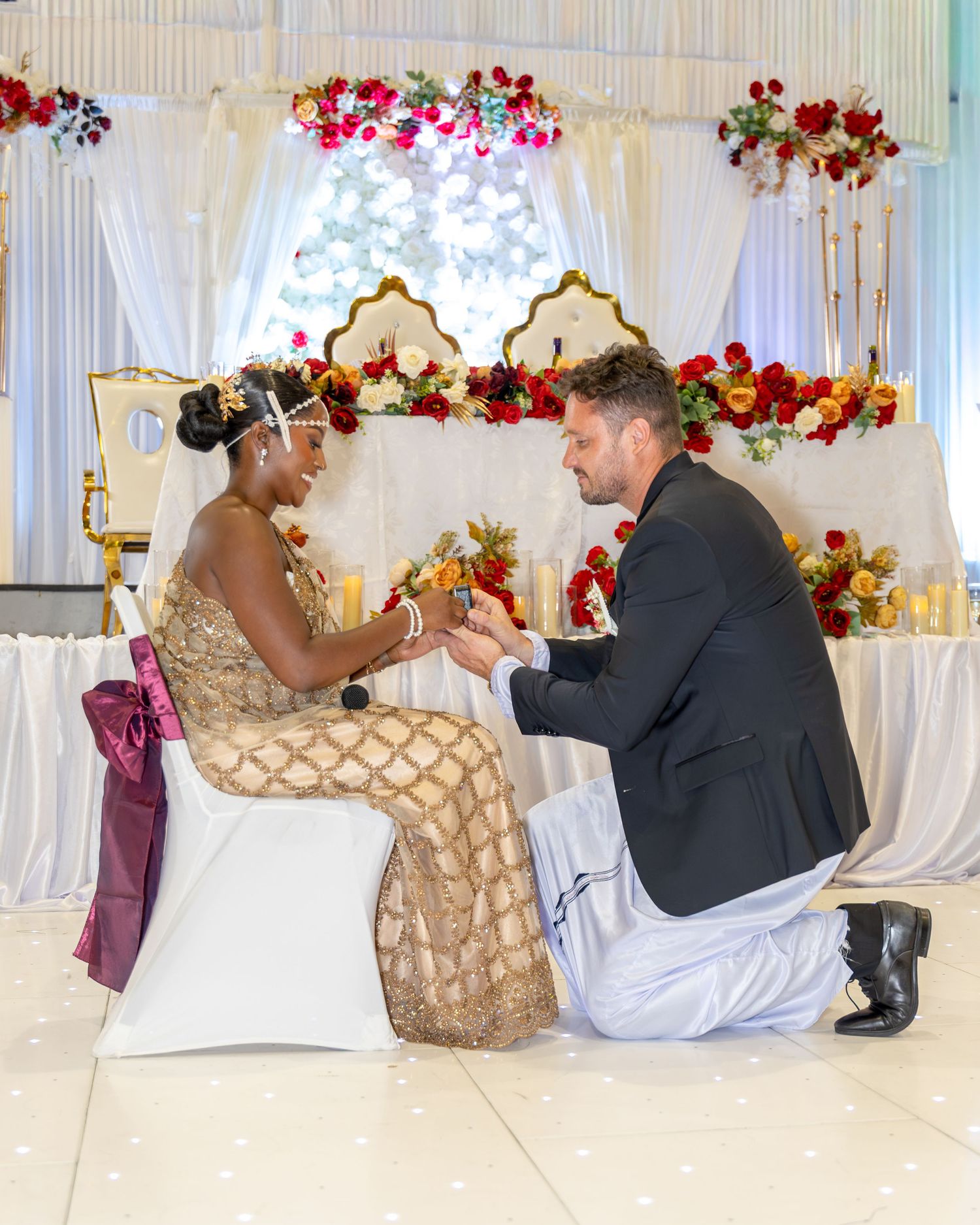 Man kneeling before bride in gold traditional African dress at a wedding ceremony with floral backdrop.