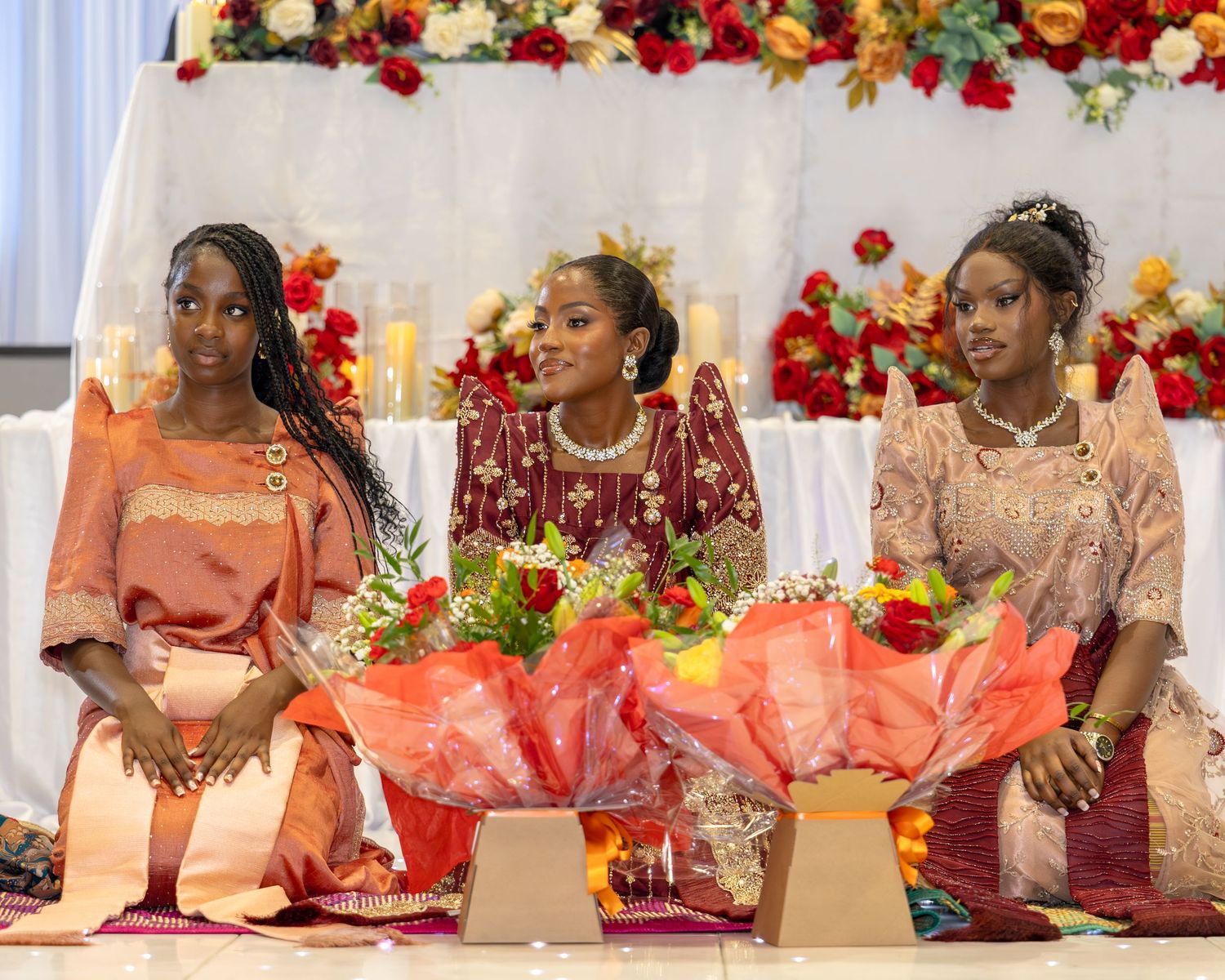 Three women in elegant traditional African dresses seated with bouquets at a wedding reception.